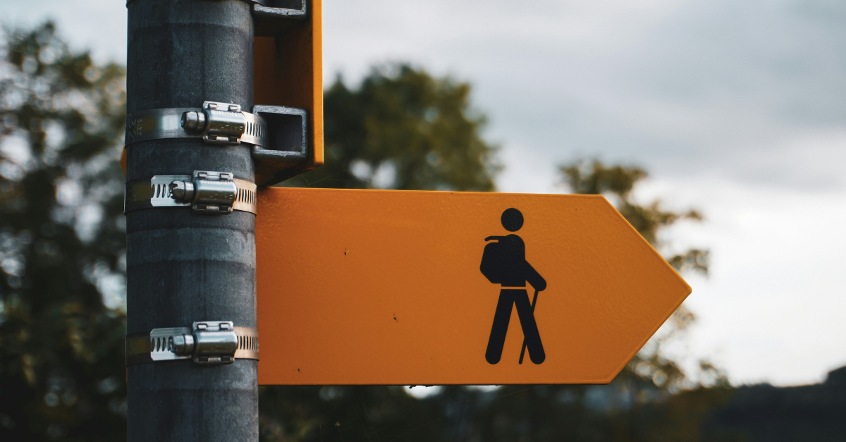 Yellow trail sign with a walking figure and backpack, mounted on a pole, pointing right against a blurred background of trees and overcast sky.