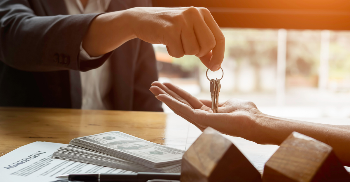 A real estate professional hands over keys to a client across a table with money and an agreement, suggesting a successful real estate deal.