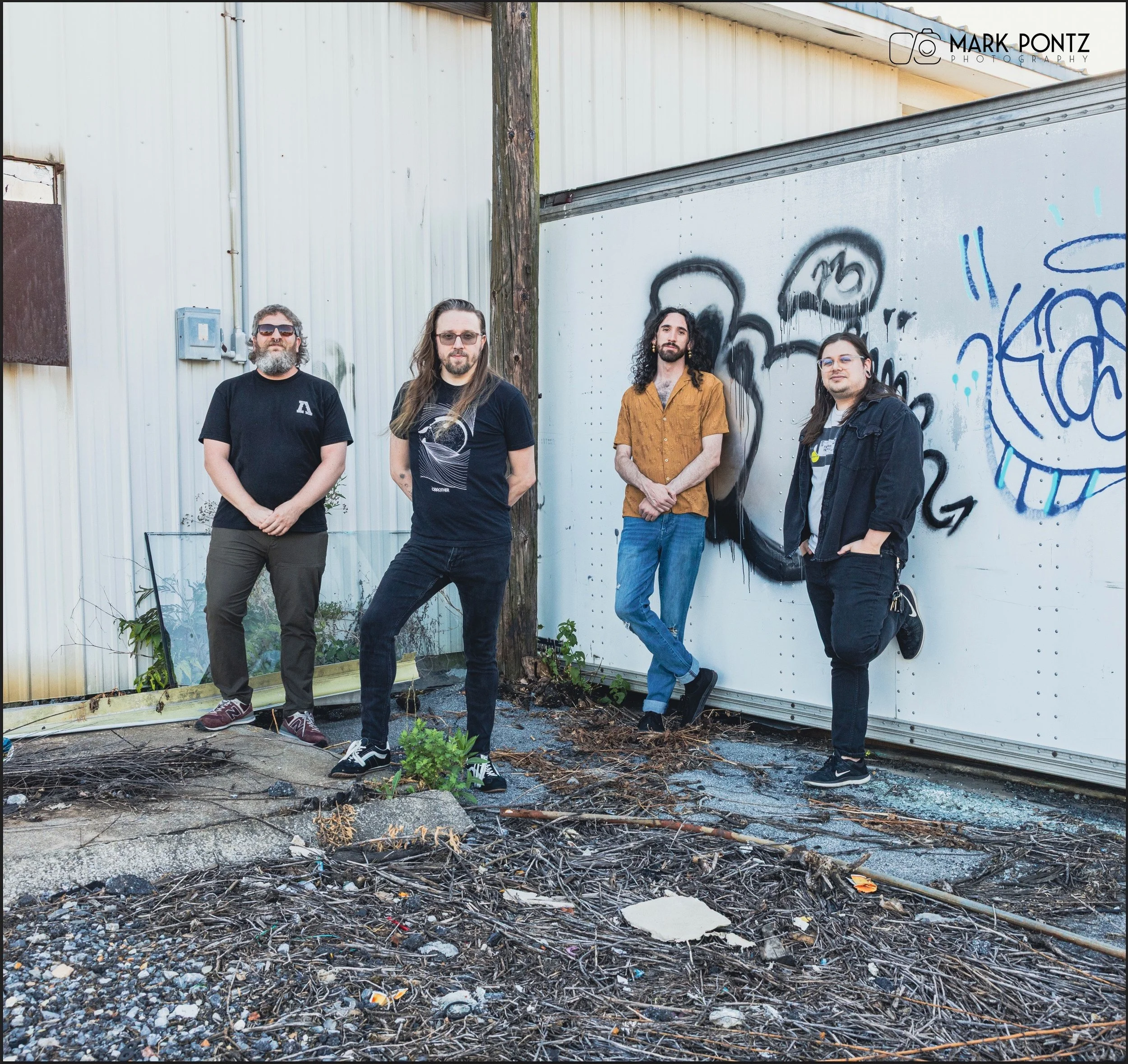 Four men standing outdoors in front of a graffiti-covered wall and a weathered metal building, with gravel and debris on the ground.