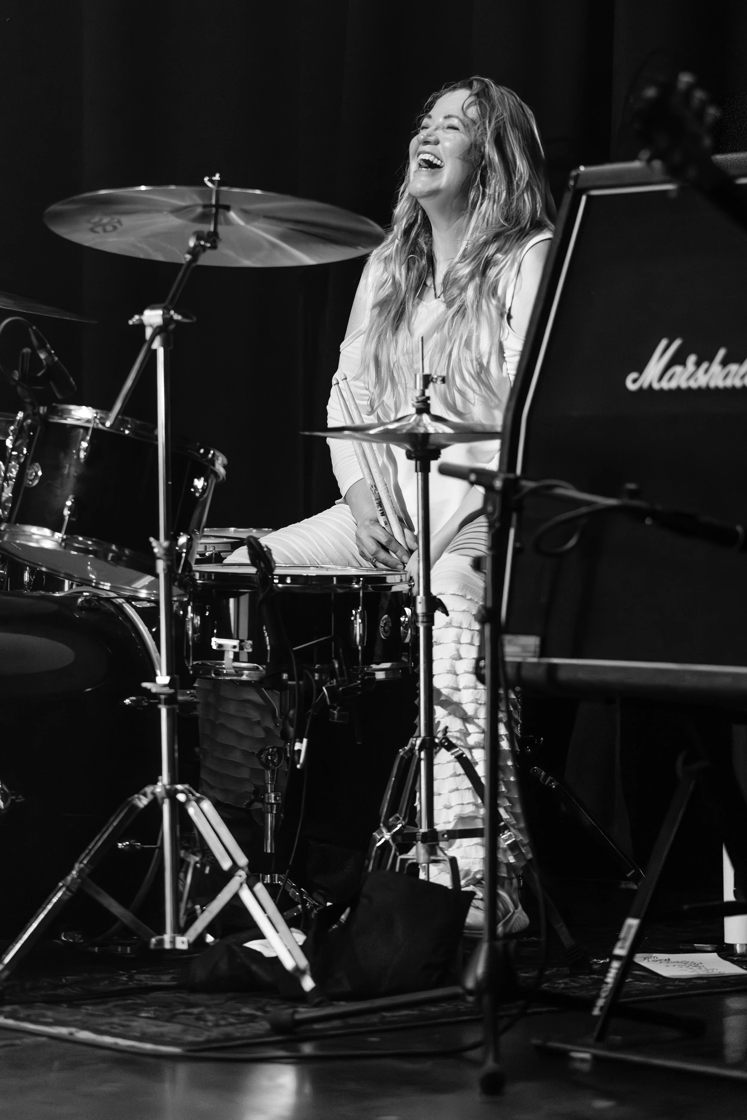 A woman playing drums and smiling joyfully during a performance, set on a stage with musical equipment and instruments.
