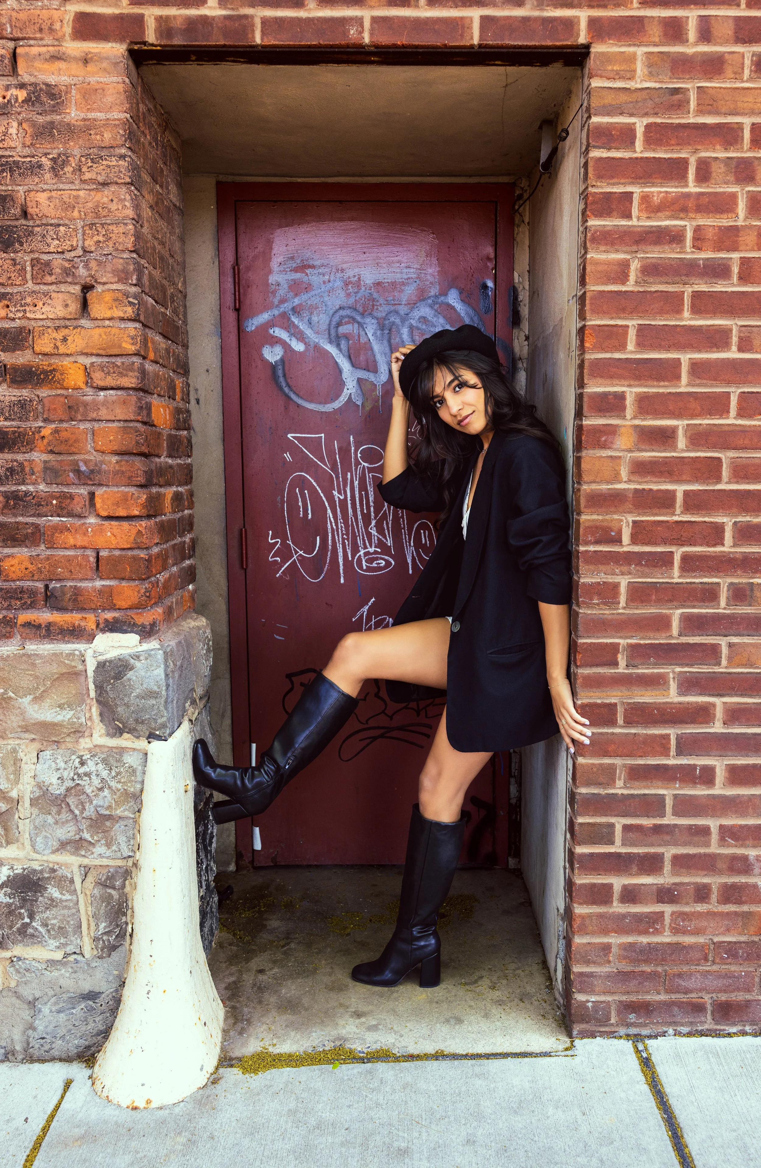 A woman wearing a black coat, black hat, and black knee-high boots posing in a brick alleyway with a graffiti-covered door behind her.