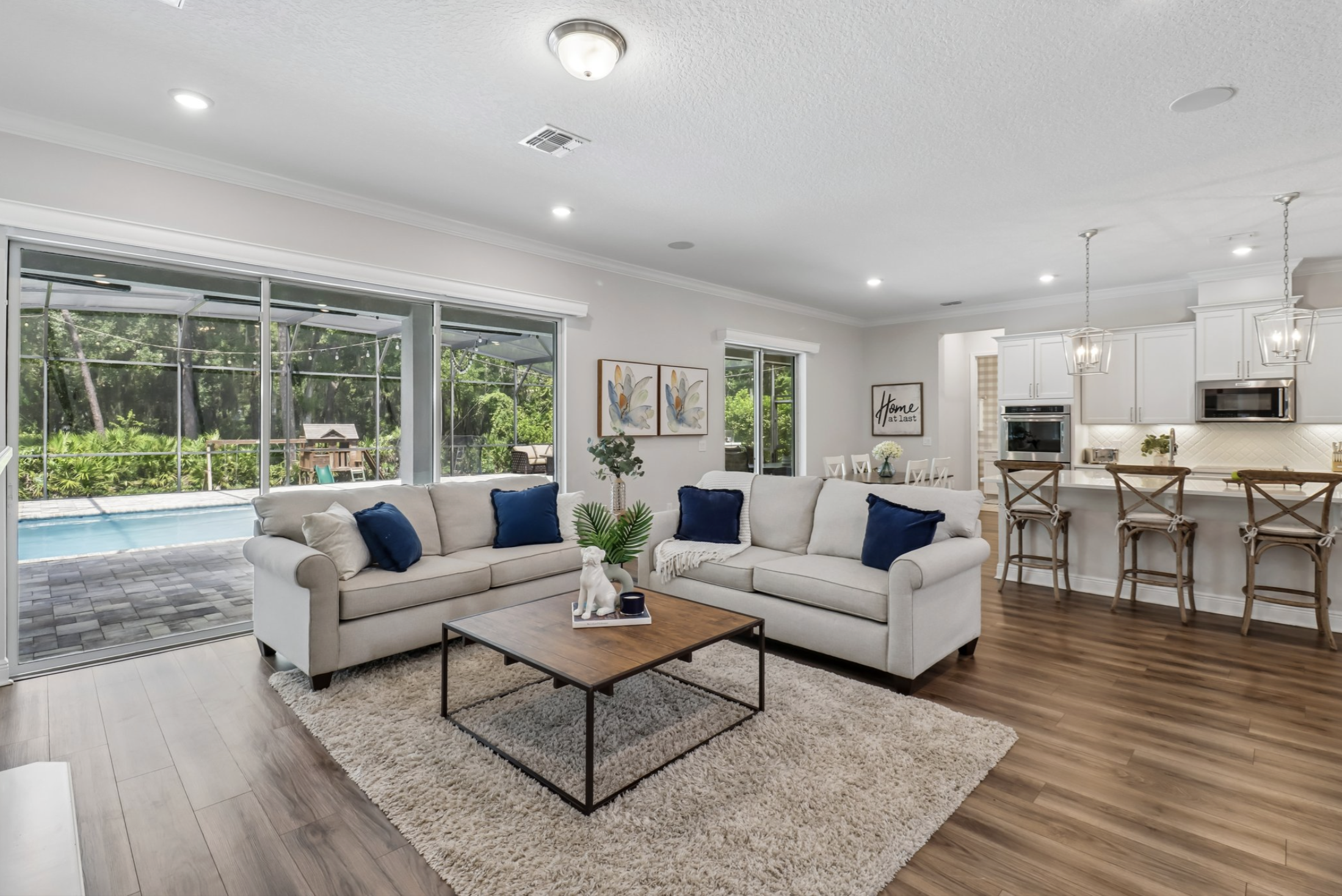 Living room with a beige sofa, blue pillows, a wooden coffee table, and sliding glass doors leading to an outdoor area with a pool and greenery