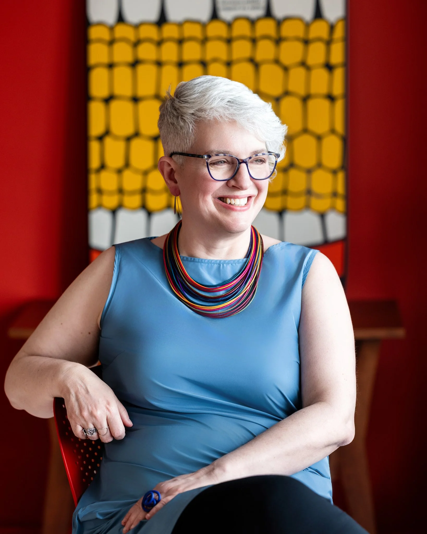 Bright and colorful headshot of woman with short hair sitting in front of red and yellow artwork