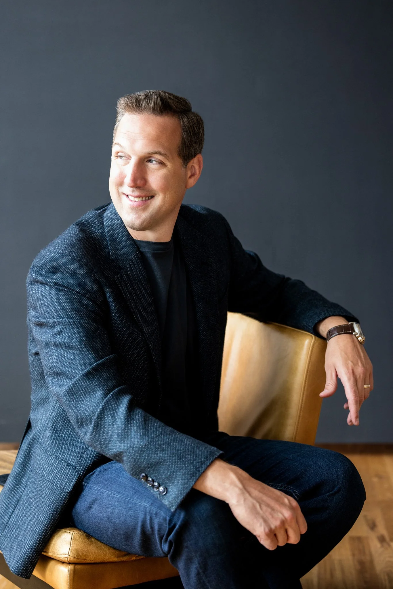 Casual professional headshot of a man on a leather chair with blue background