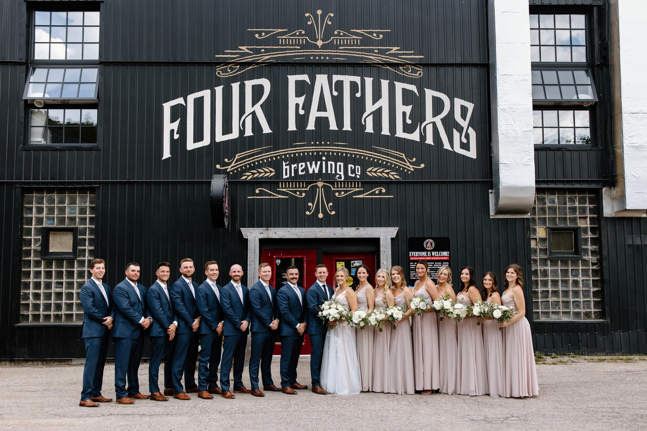 bride and groom posing outside cambridge wedding venue four fathers brewing co. with their wedding party