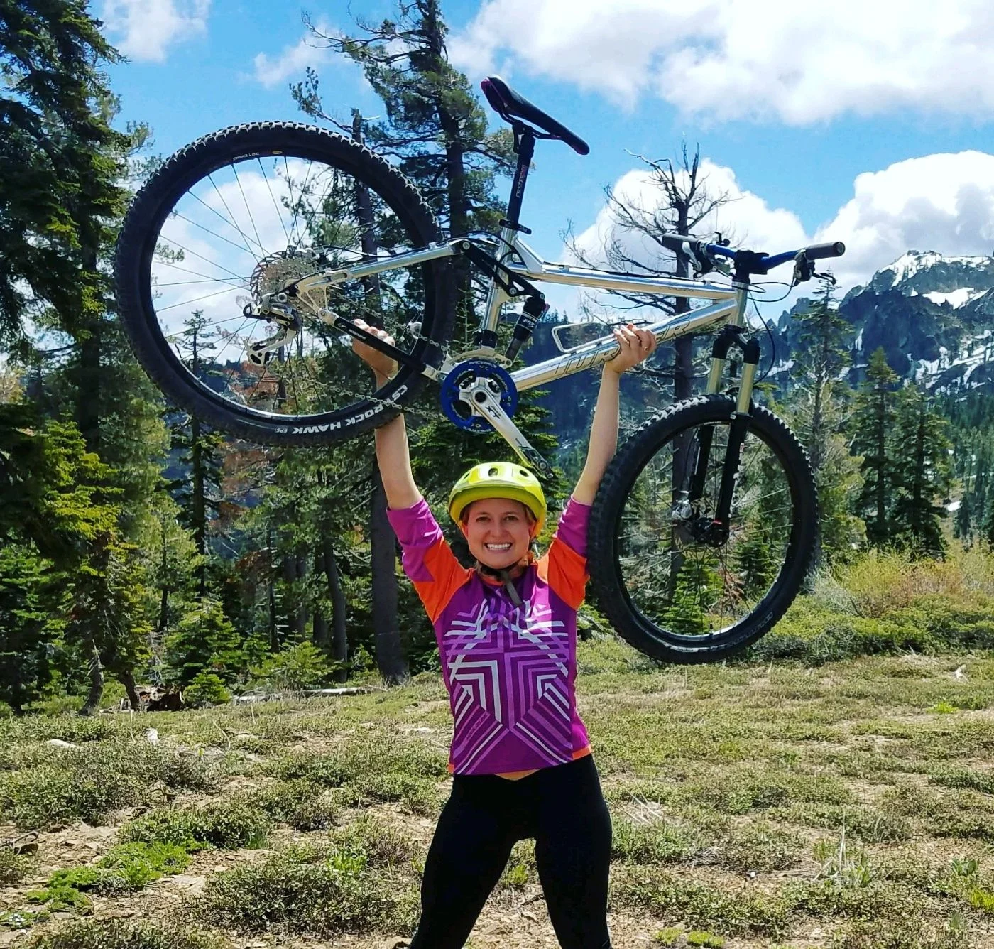 a white women wearing a purple cycling jersey with red short sleeves and black shorts smiles at the camera as she lifts her white mountain bike over her head. there are pine trees and snow covered mountains in the background.