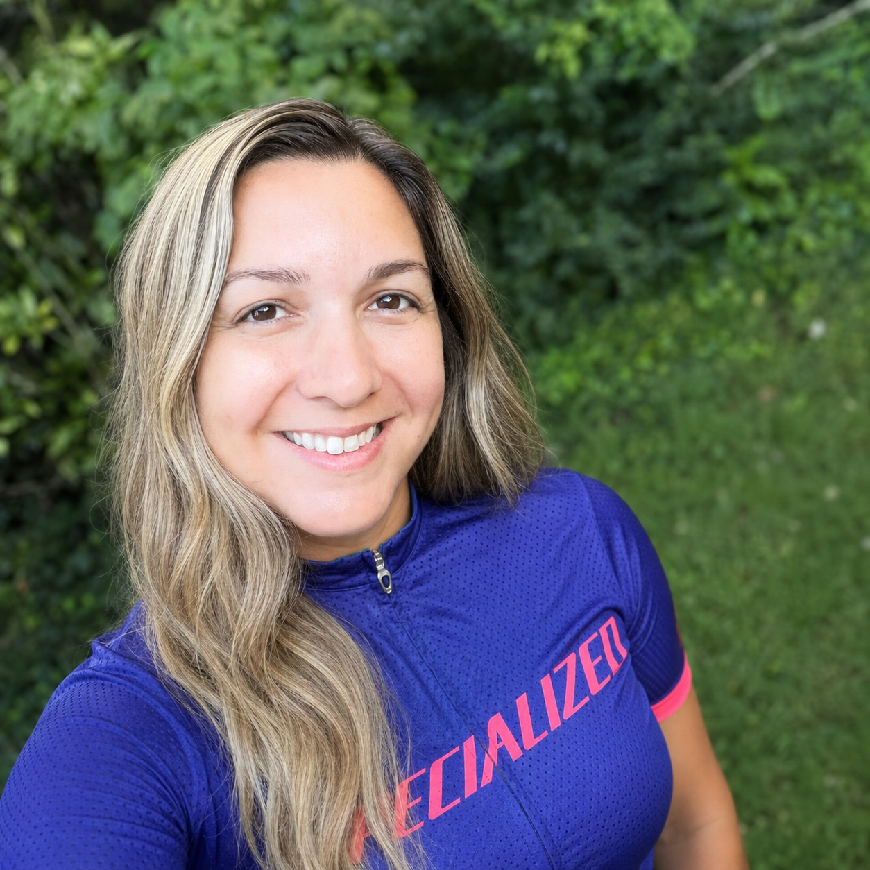 a woman with long blonde hair smiles at the camera. she's standing in front of green bushes and is wearing a blue bike jersey with pink letters on it.