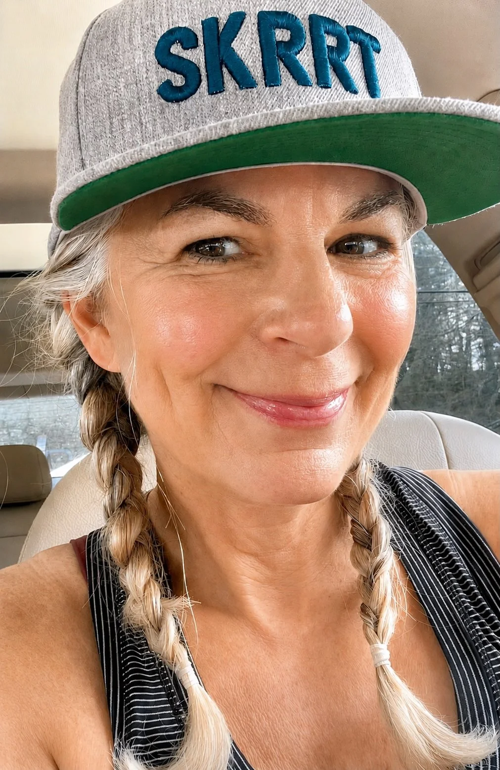 Becky, a white woman with greying light brown hair in two braids wearing a grey brimmed cap with dark blue SKRRT embroidered across it smiles at the camera.