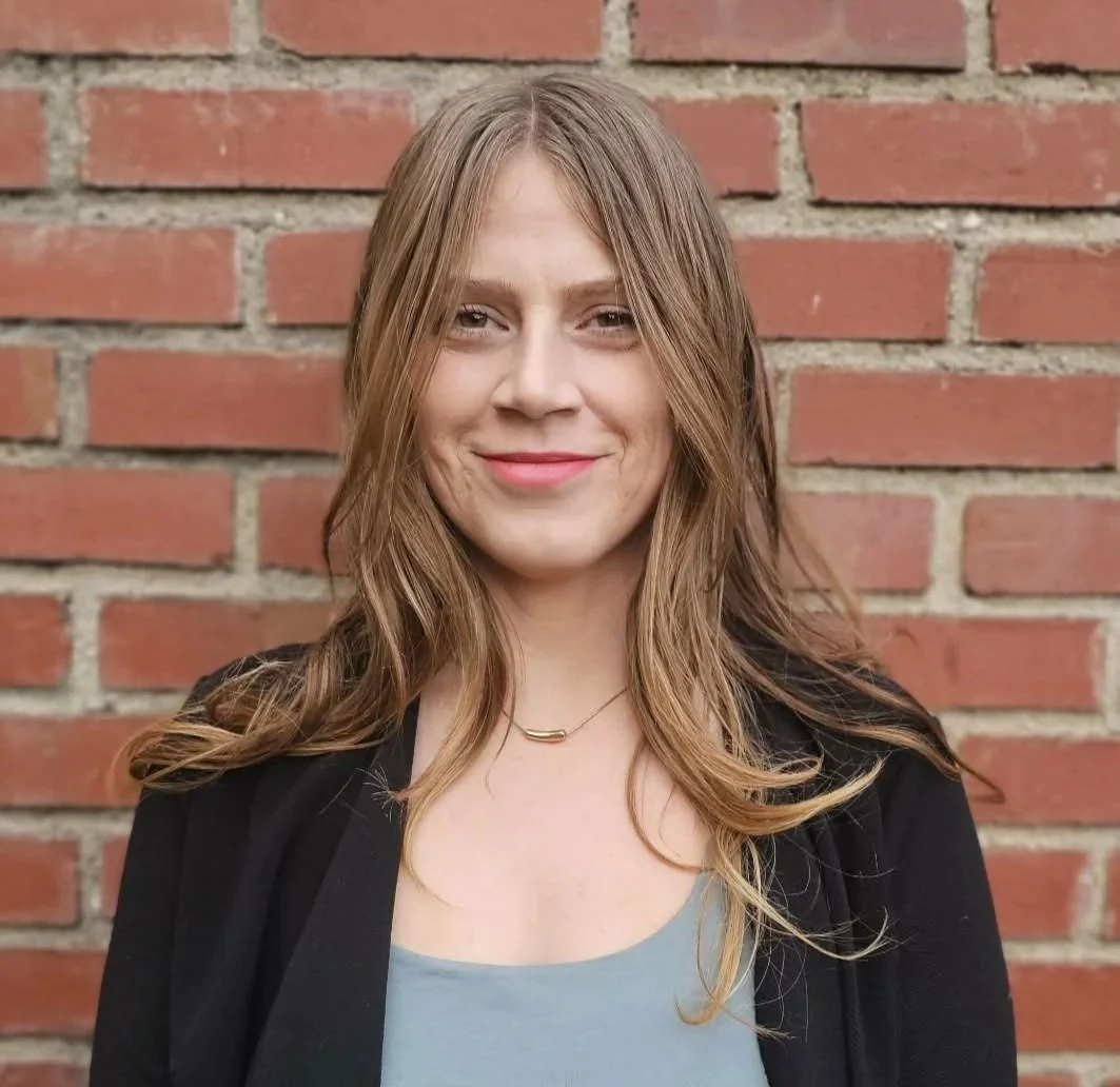 a woman with long light brown hair parted in the middle smiles at the camera. she's standing in front of a red brick wall and is wearing a blue grey shirt and a black sweater.