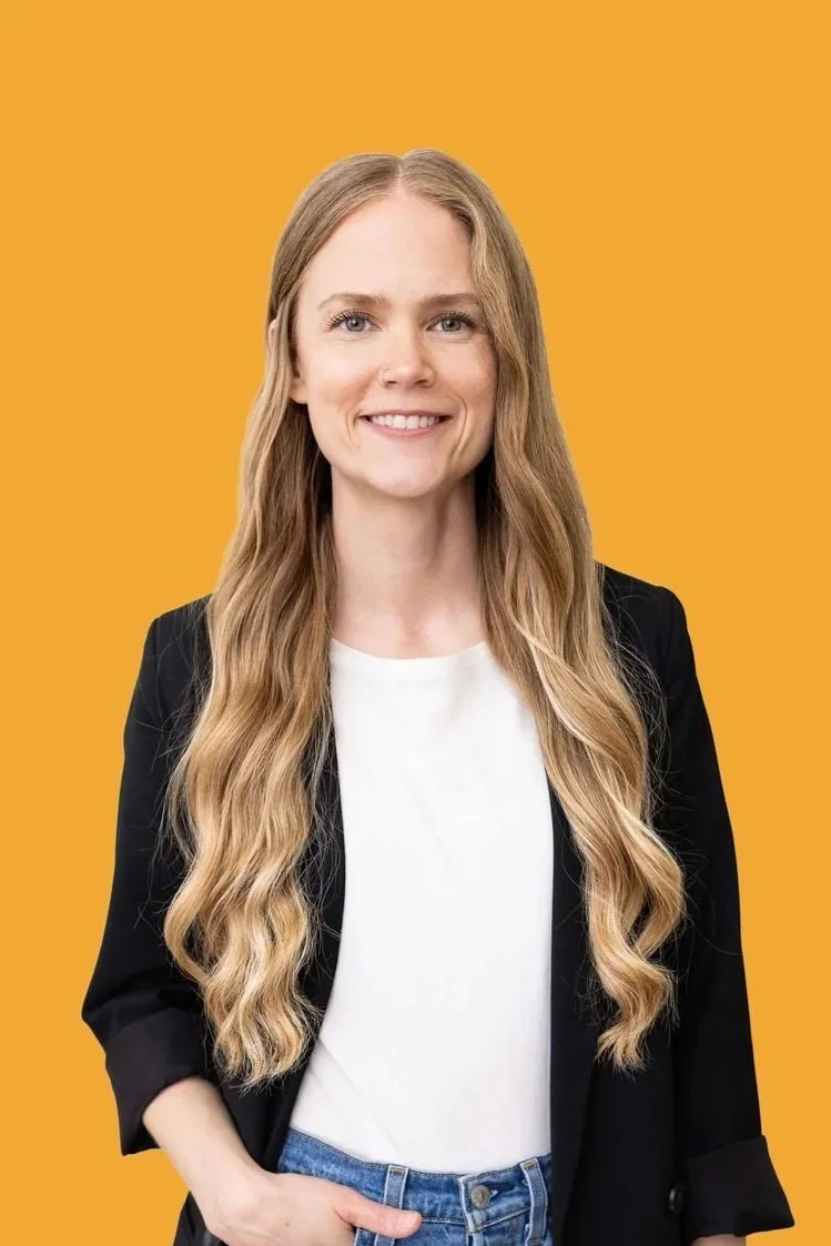 A woman with long wavy blond hair wearing a black blazer over a white T-shirt, smiling, against a yellow background.