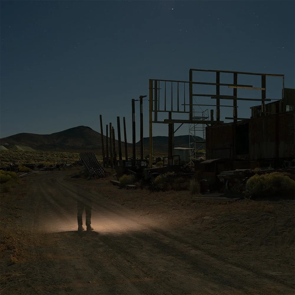 Nighttime scene of a dirt road with a person faintly illuminated, leading past old, abandoned wooden structures and a landscape of hills or mountains under a starry sky.