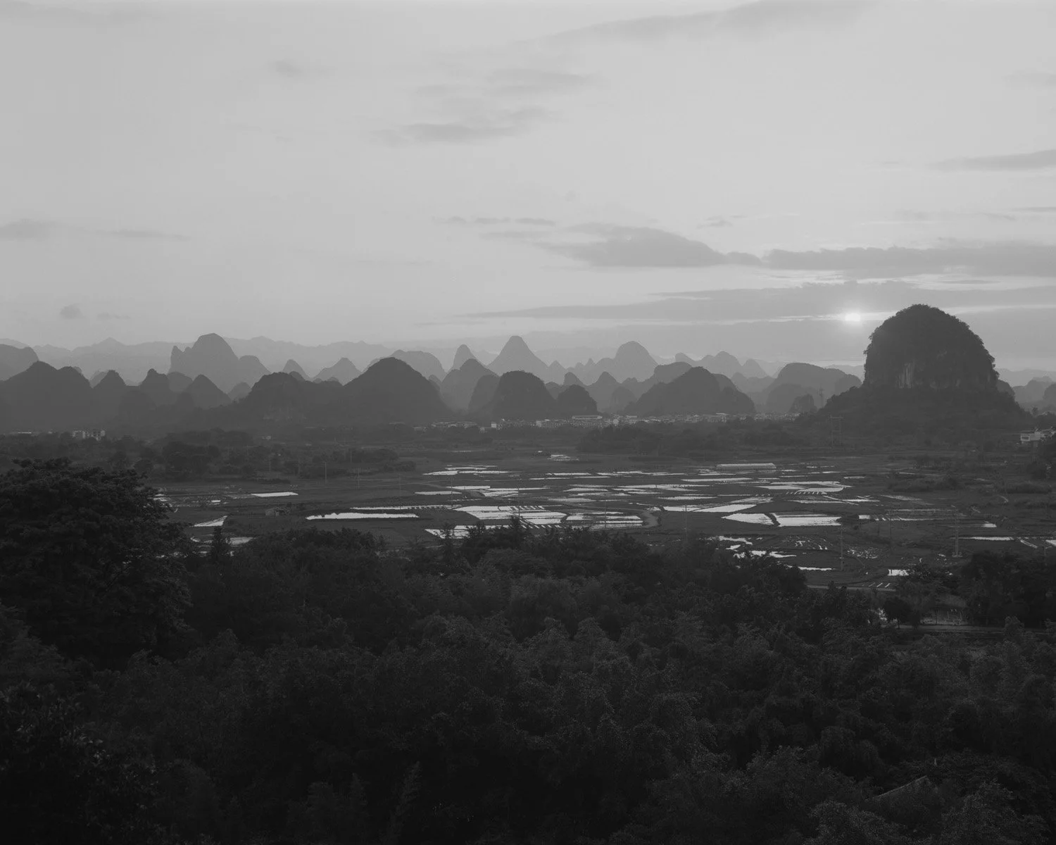 Black and white landscape featuring distant mountain ranges, fields with water reflections, and a large hill on the right side under a sky with clouds and a setting or rising sun.