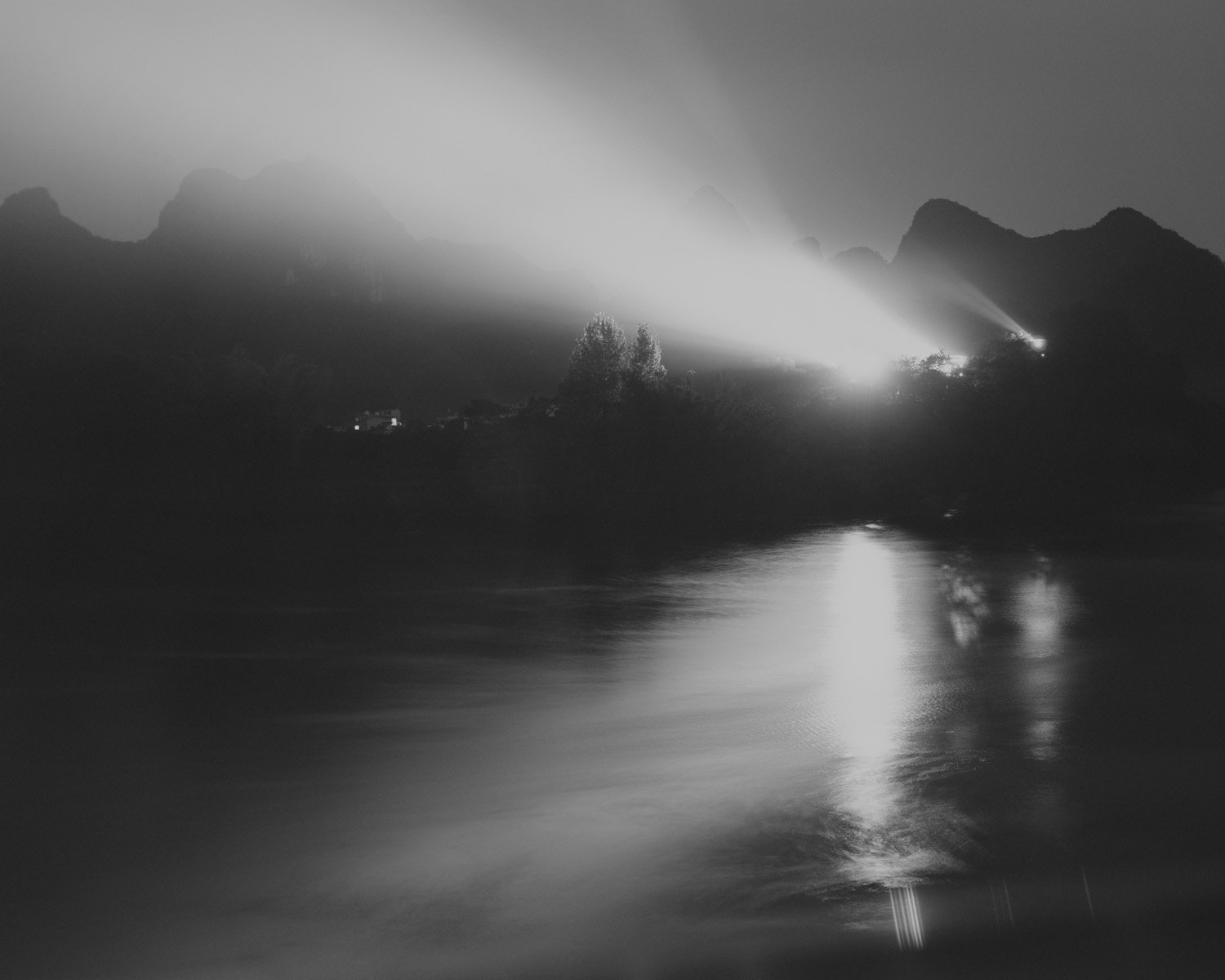 Black and white photo of a river with mountains in the background, illuminated by a bright light source on the right, creating reflections on the water.