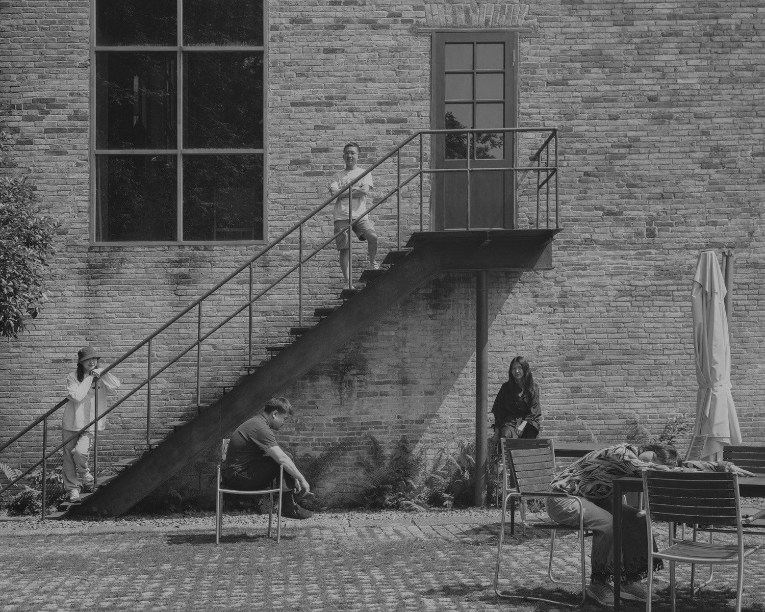 Group of five people relaxing outside near a brick building with large window and glass door. One person is sitting on a bench, two others are sitting on the ground, and two are on the staircase, one standing on the stairs and another sitting on a le