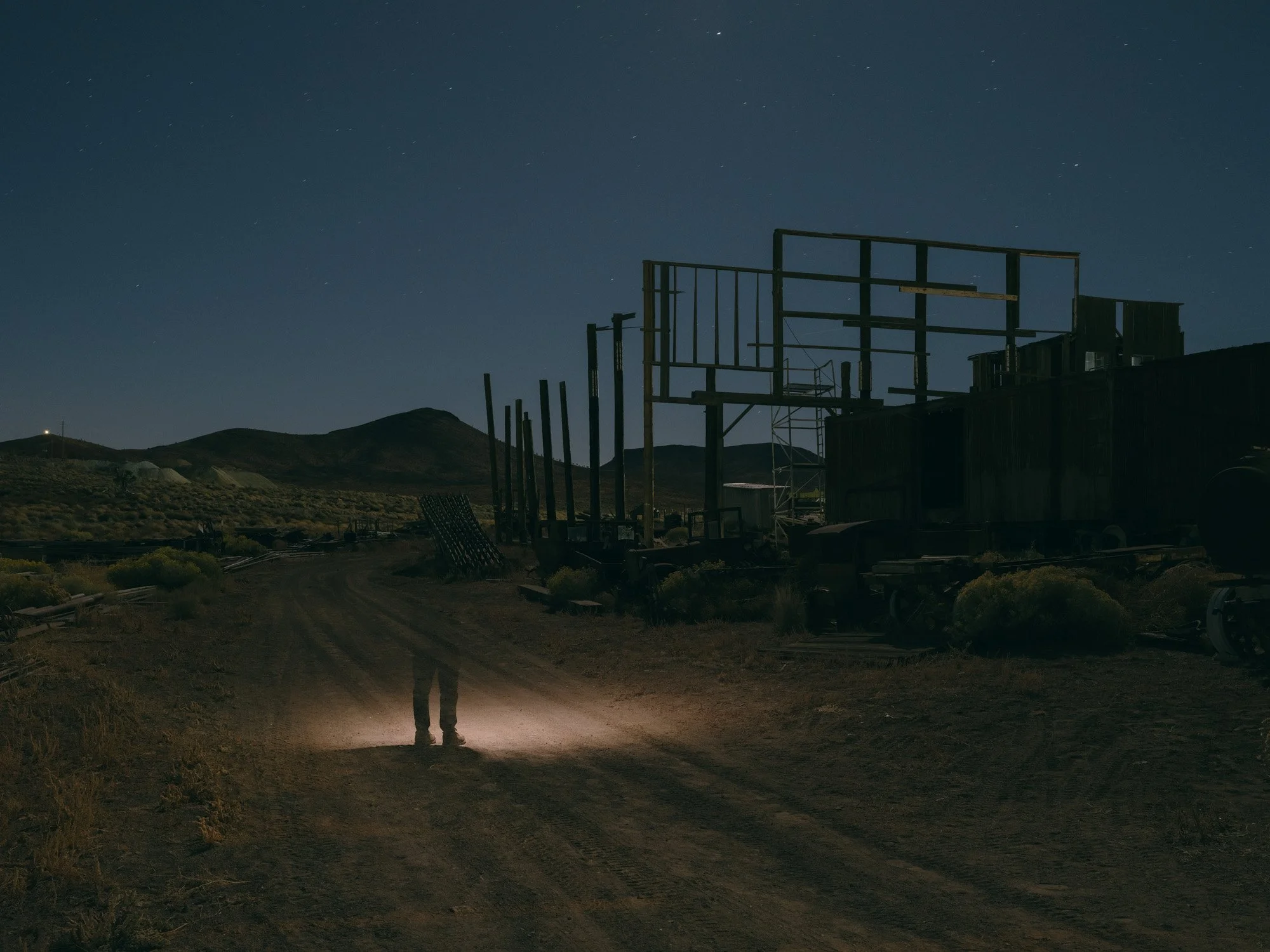Visual art by Beihua Guo exploring historical erasure. A nighttime color photo of a spectral figure standing before the abandoned wooden structures of the Goldfield, Nevada railroad yard.