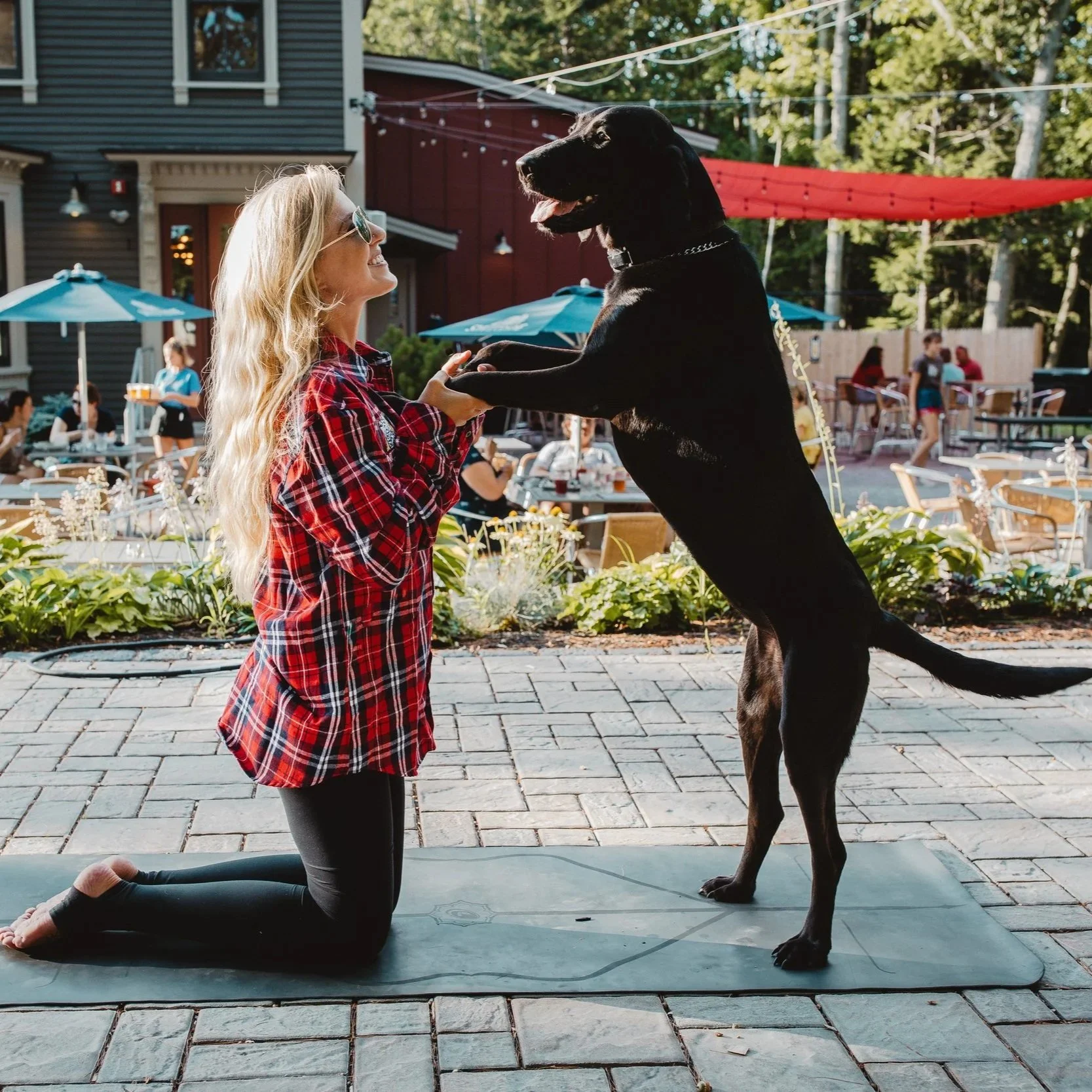 A woman kneeling on a yoga mat, holding hands with a large black dog that is standing on its hind legs outdoors at a restaurant patio.