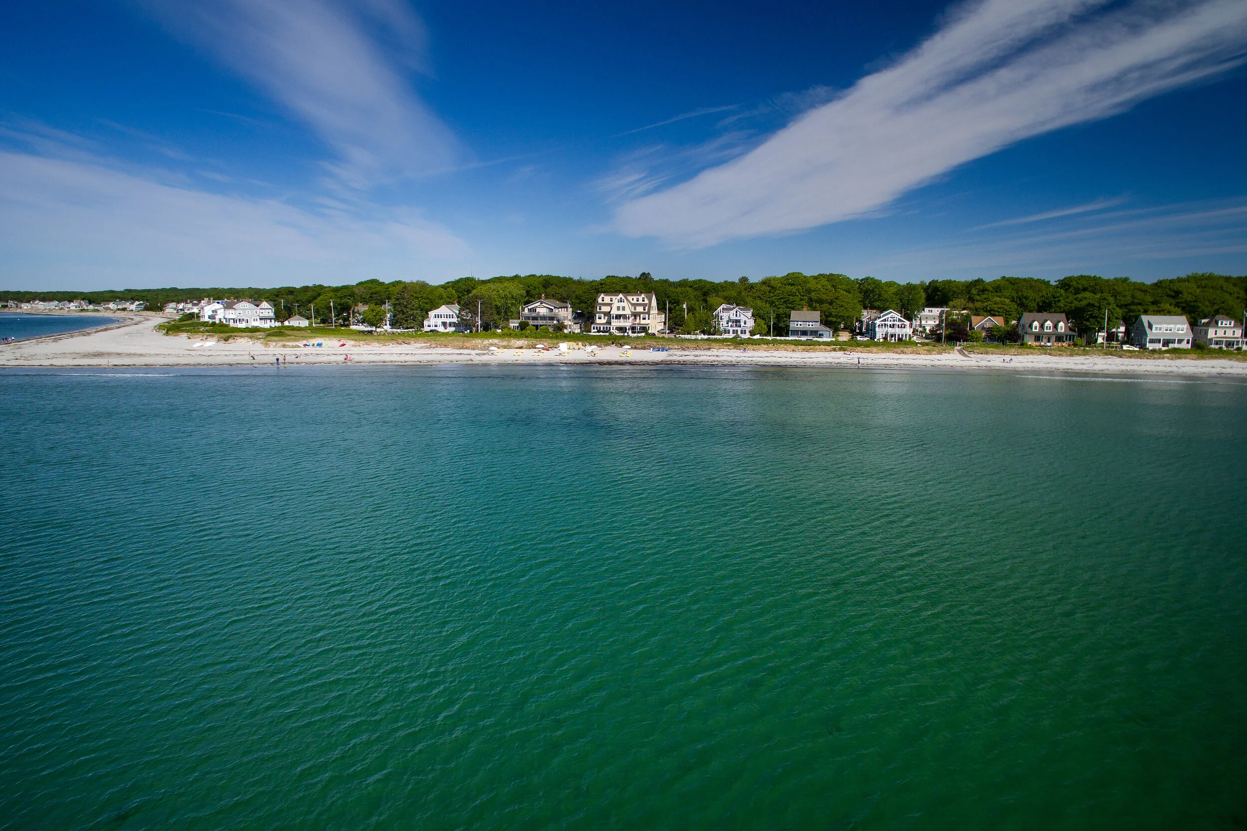 Coastal neighborhood with beachfront houses and green trees under a blue sky with wispy clouds, seen across calm green water.