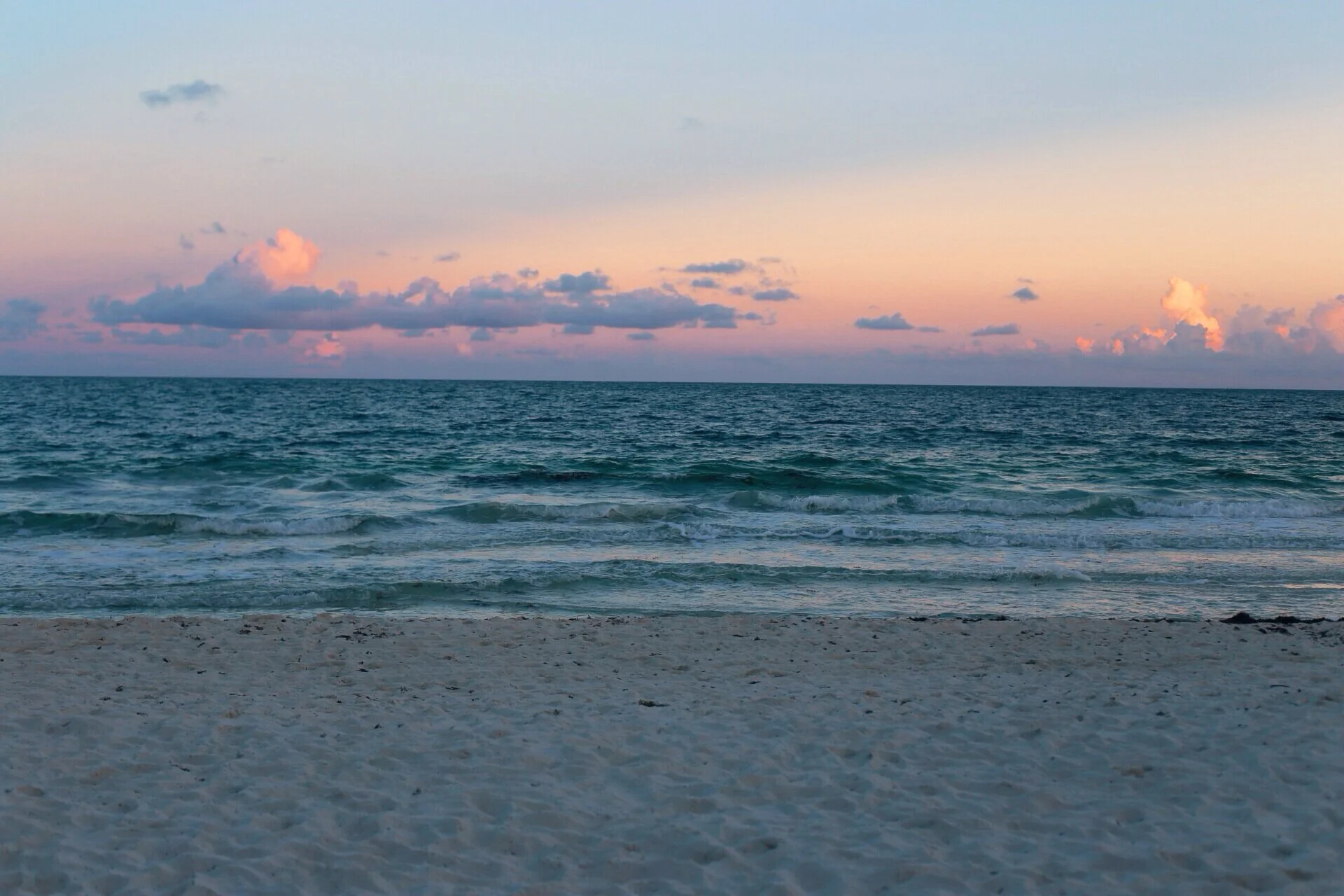 Empty sandy beach with gentle waves and a pastel-colored sky with clouds during sunset.