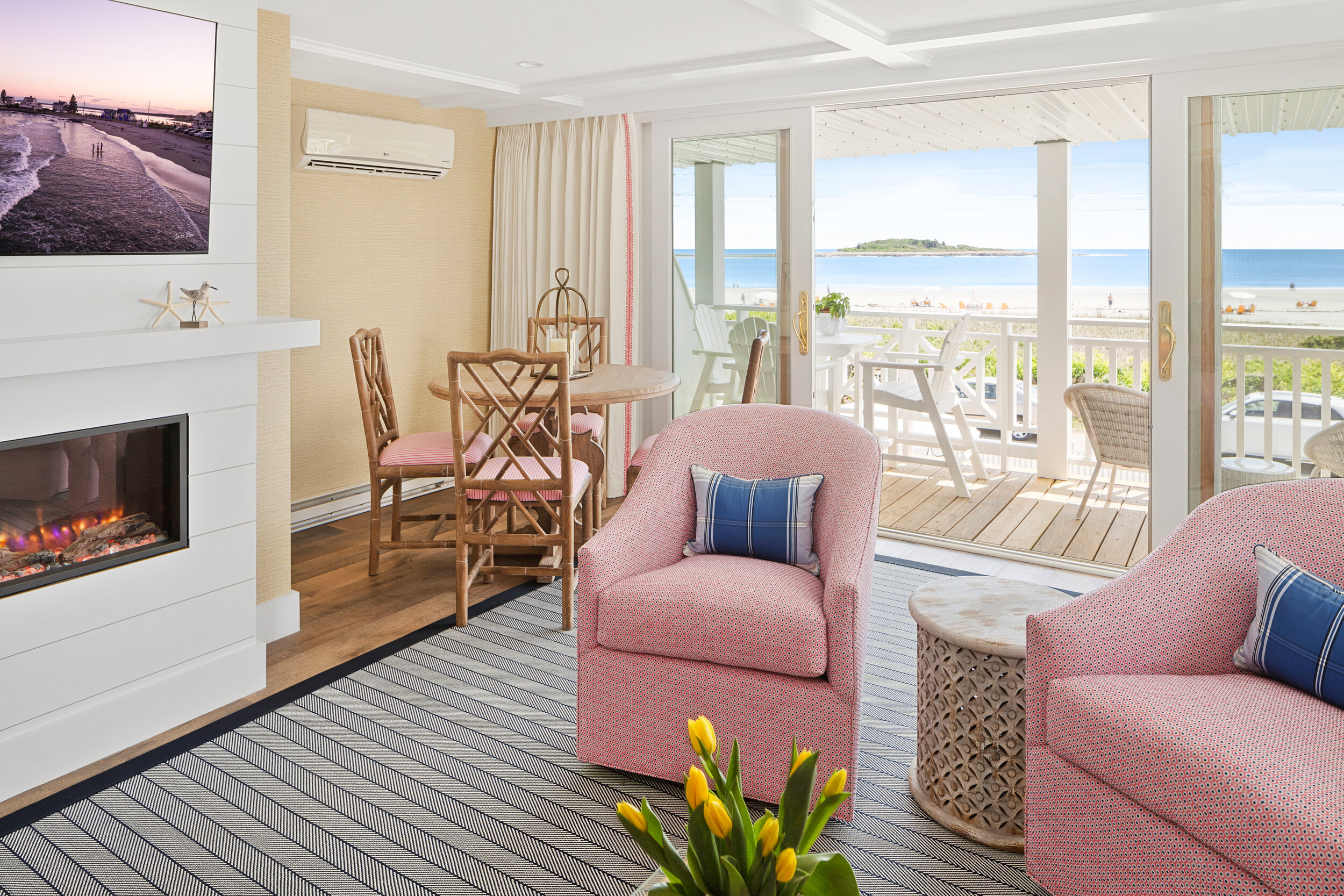 Living room with pink armchairs, a fireplace, a striped rug, a dining table with chairs, and sliding glass doors leading to a balcony overlooking the beach and ocean.