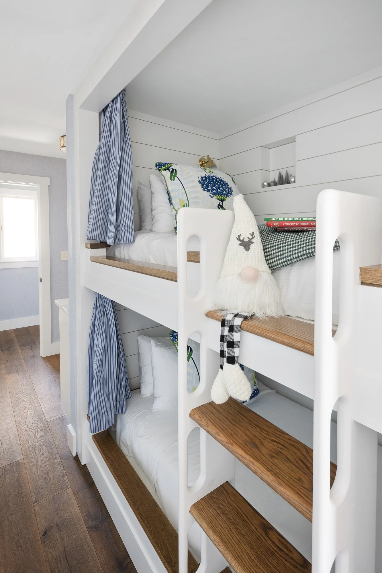 Bunk beds with white frames, wooden steps, and blue-striped curtains, decorated with holiday-themed pillows and a plush Santa gnome, in a cozy bedroom with light-colored walls and a window.