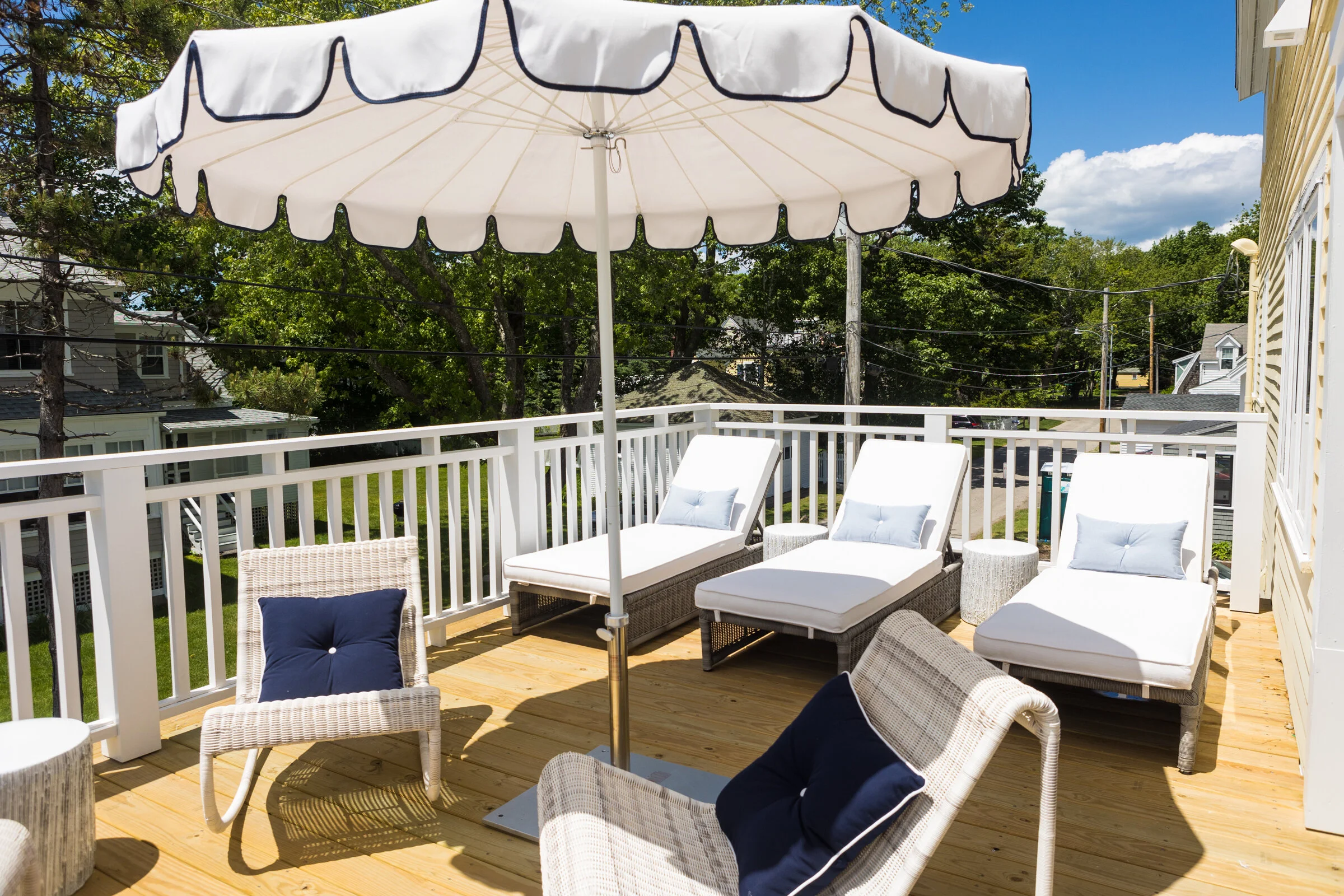 Outdoor wooden deck with white and beige outdoor furniture, including lounge chairs with light blue pillows and woven chairs with navy pillows, under a large white patio umbrella. The deck is fenced with white railing, with green trees and houses in 