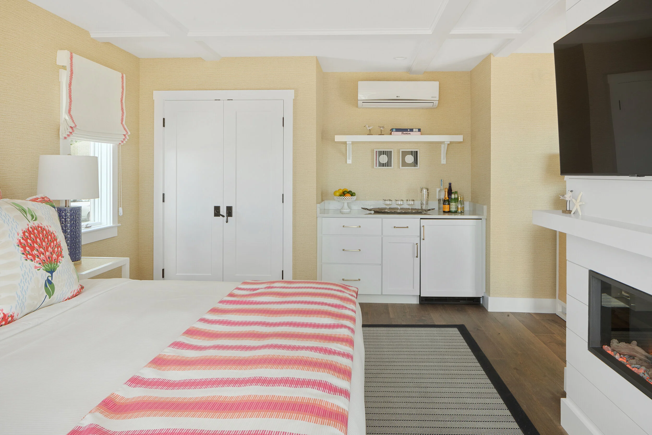 Interior of a cozy bedroom with beige walls, a large bed with floral and striped pillows, a bedside lamp, a small kitchenette with white cabinets, a countertop, a stove, and bottles, a wall-mounted air conditioning unit, a wall-mounted TV, and a fire