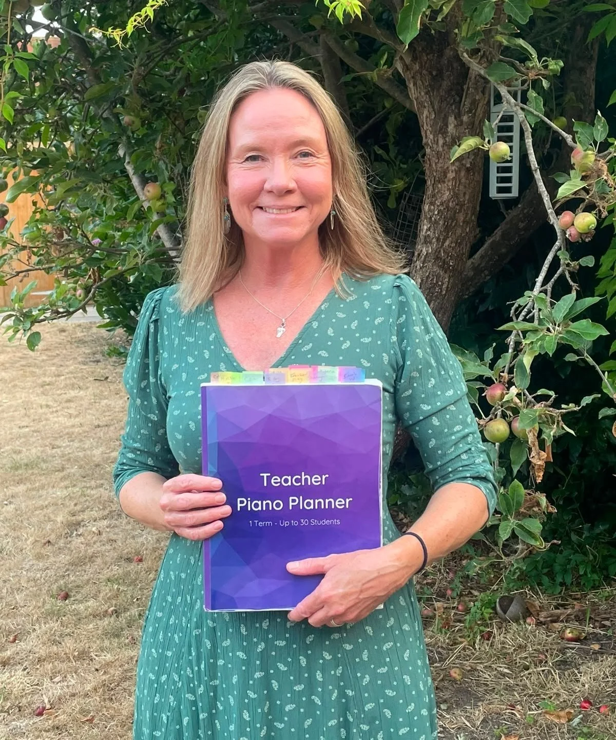 Smiling woman in a green dress standing in front of an apple tree holding a purple Teacher Piano Planner
