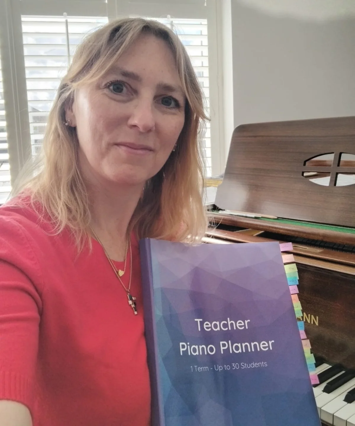 Blonde womand with a red top holding a purple Teacher Piano Planner while sat at her piano