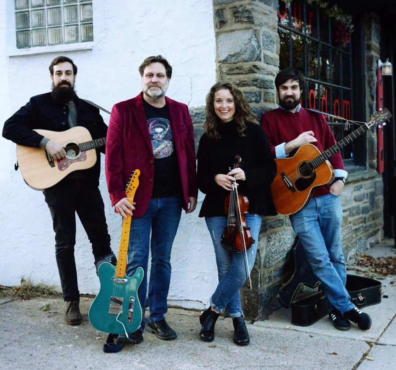 Three men hold guitars and one woman hold a violin as they all stand on a sidewalk outside a bar.