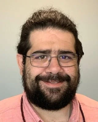 A man with glasses and a beard wearing a pink shirt and a beaded necklace, smiling at the camera.
