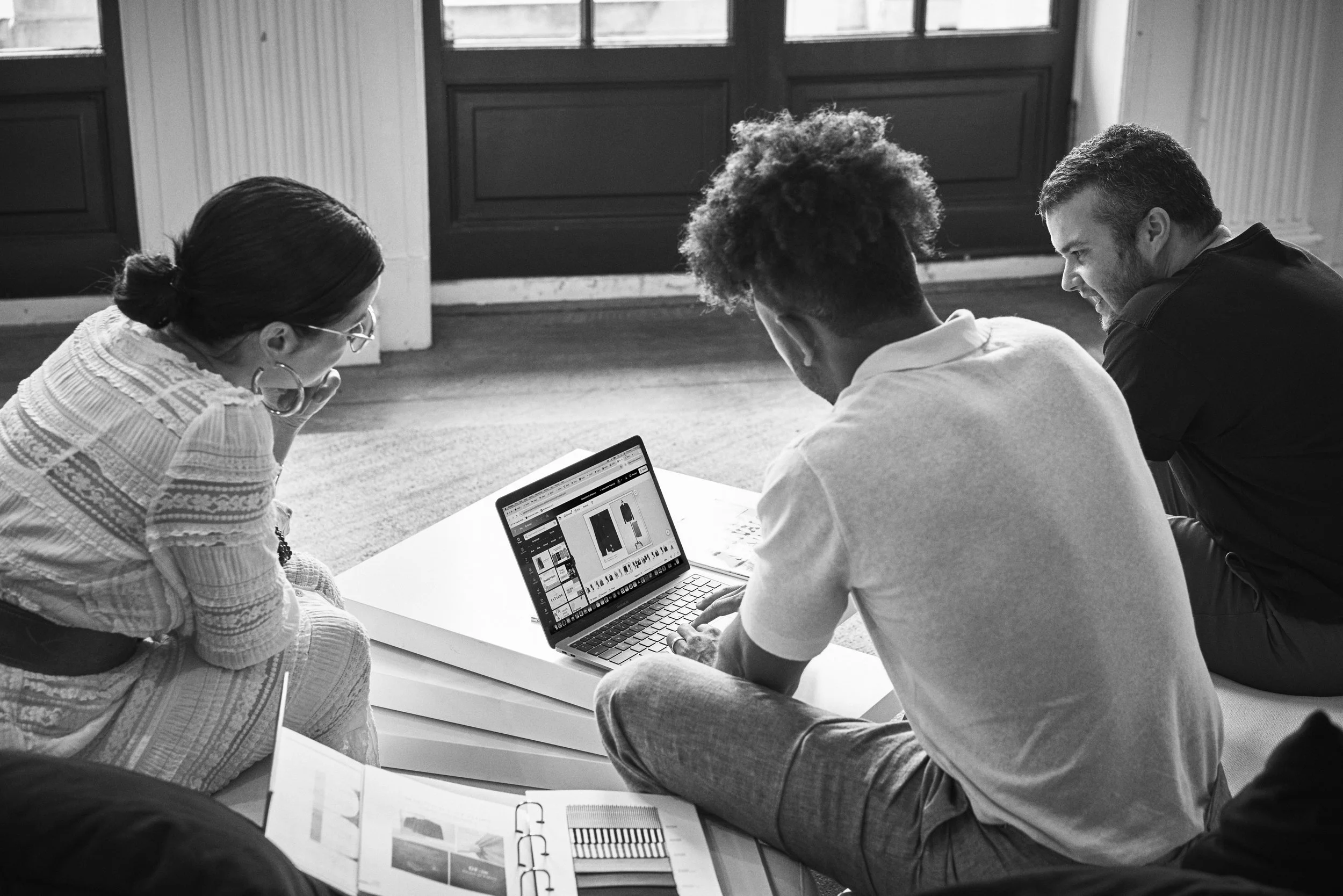 Three people sitting on the floor around a laptop, looking at the screen, in a room with large windows.