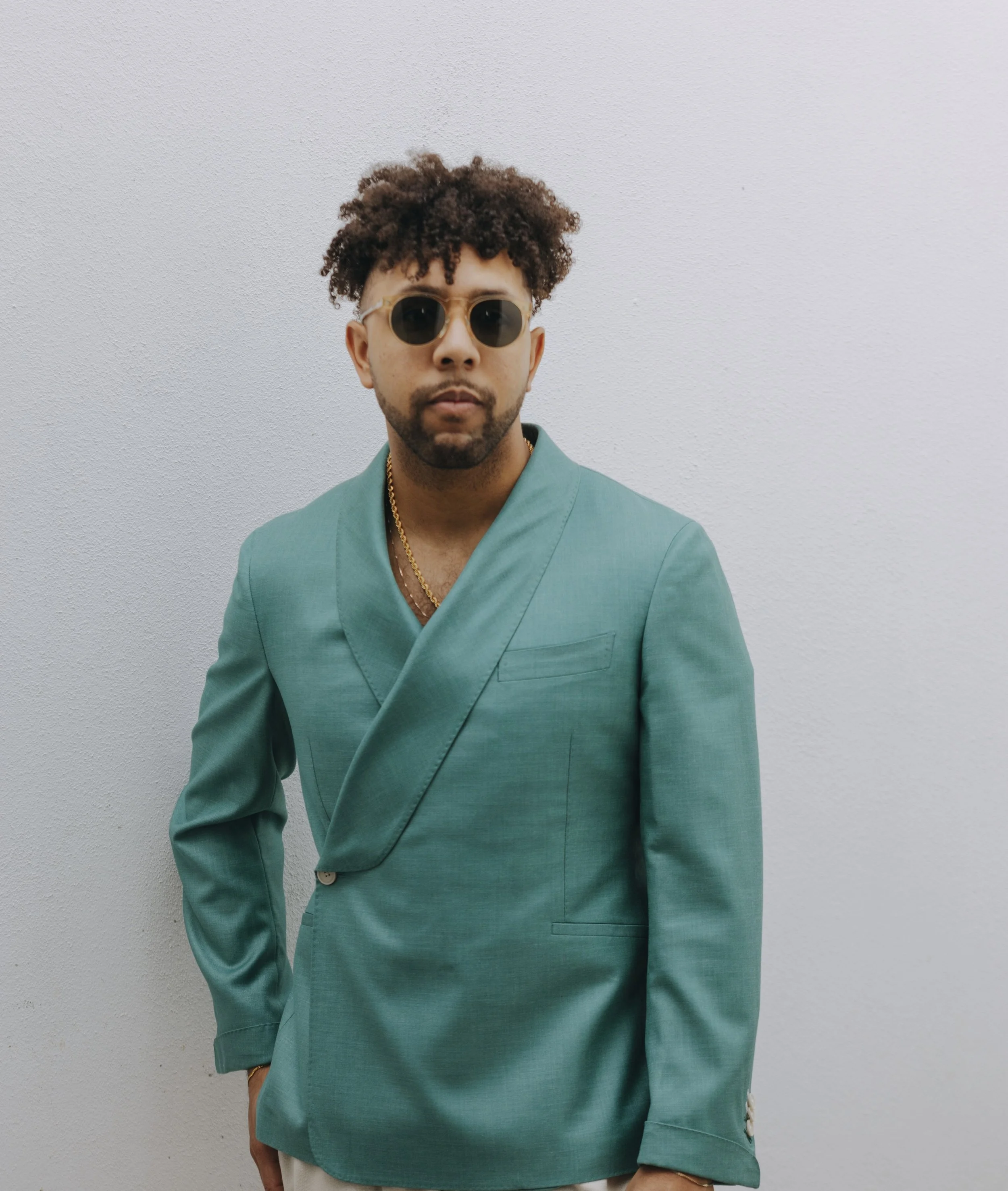 Man wearing teal suit, sunglasses, and gold chains, standing against a plain white wall.