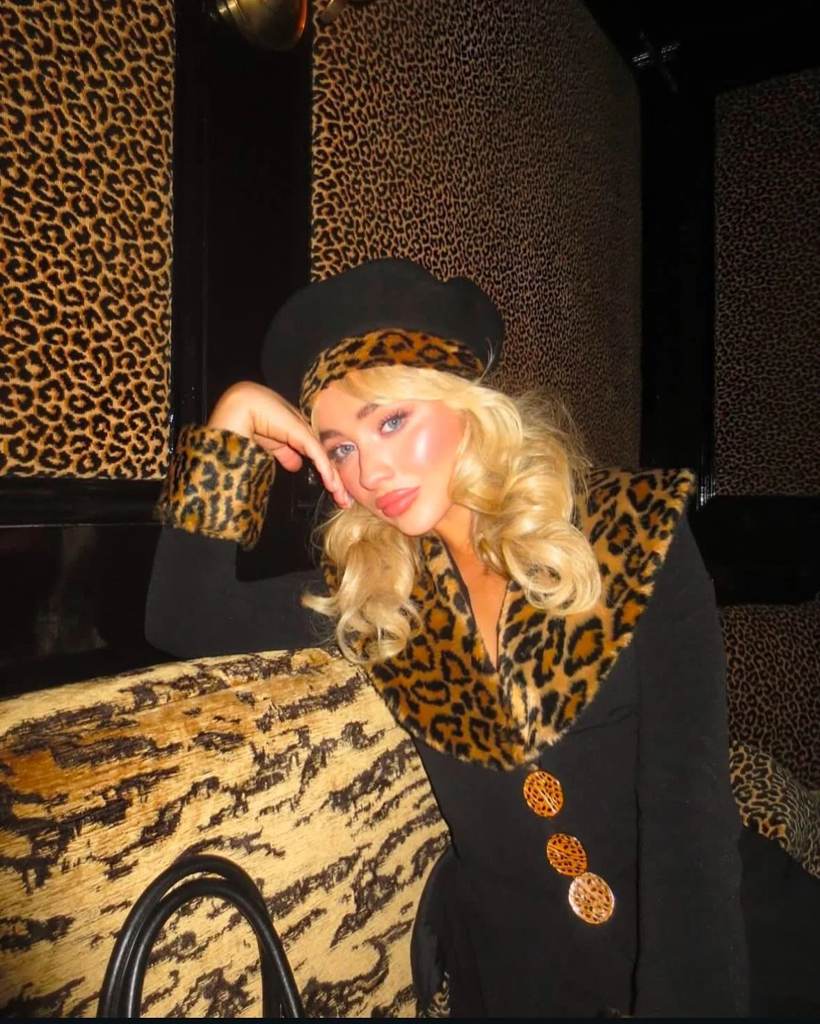 A woman with blonde curly hair wearing a black hat, black dress with leopard print buttons and leopard print accents on sleeves and collar, posing with her head resting on her hand in front of a leopard print backdrop.