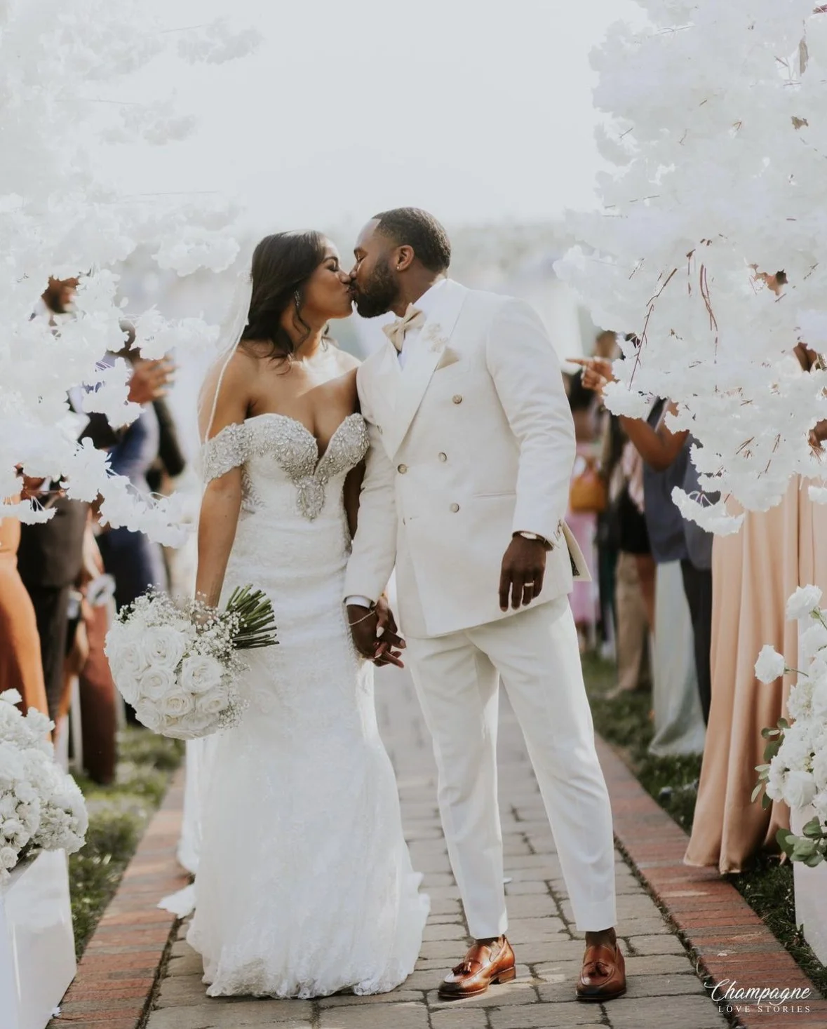 A bride and groom kiss during their wedding ceremony outdoors, surrounded by white floral arrangements and wedding guests.