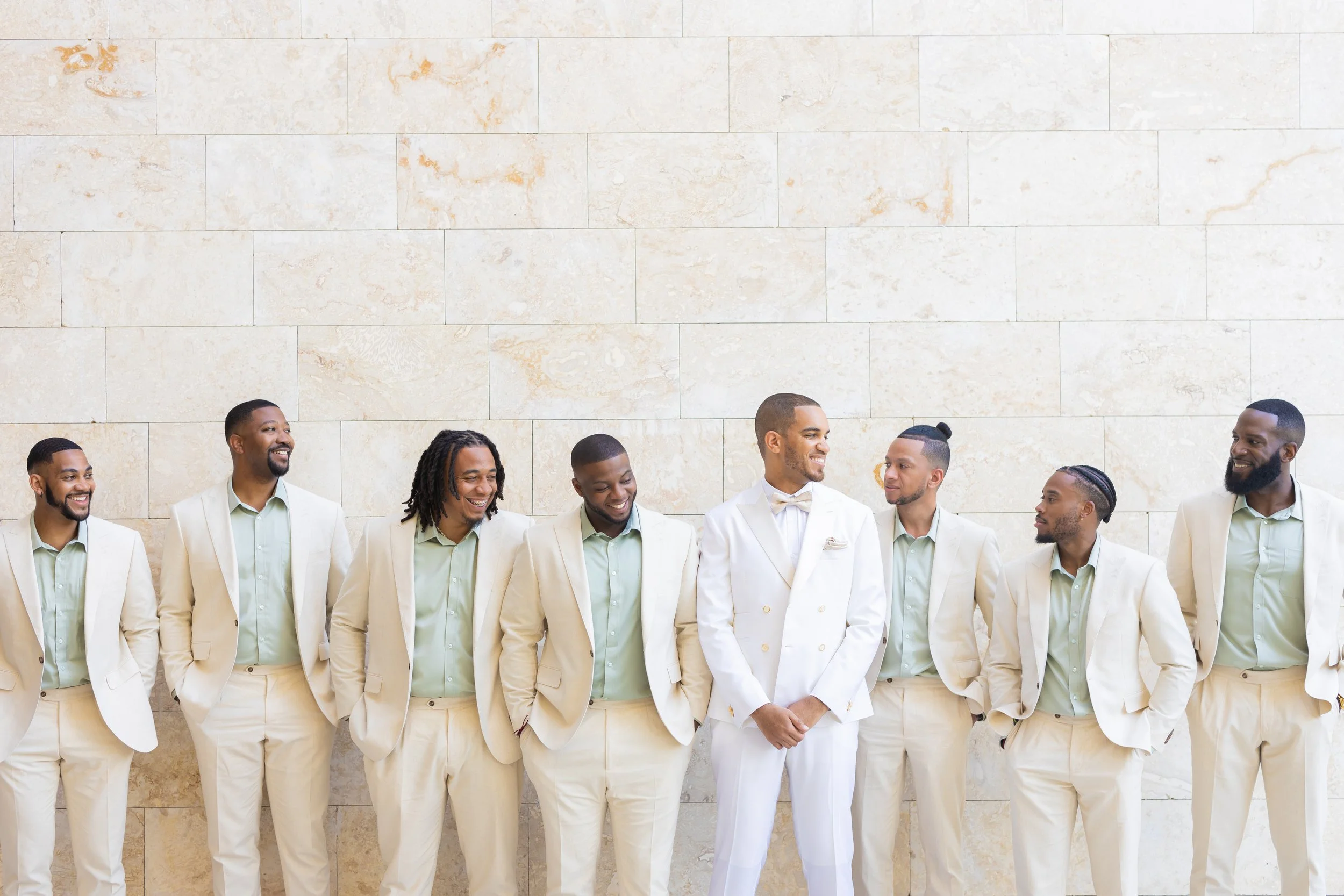 A group of eight men dressed in beige suits and light green shirts, with one man in a white tuxedo, standing against a stone wall, smiling and looking at each other.