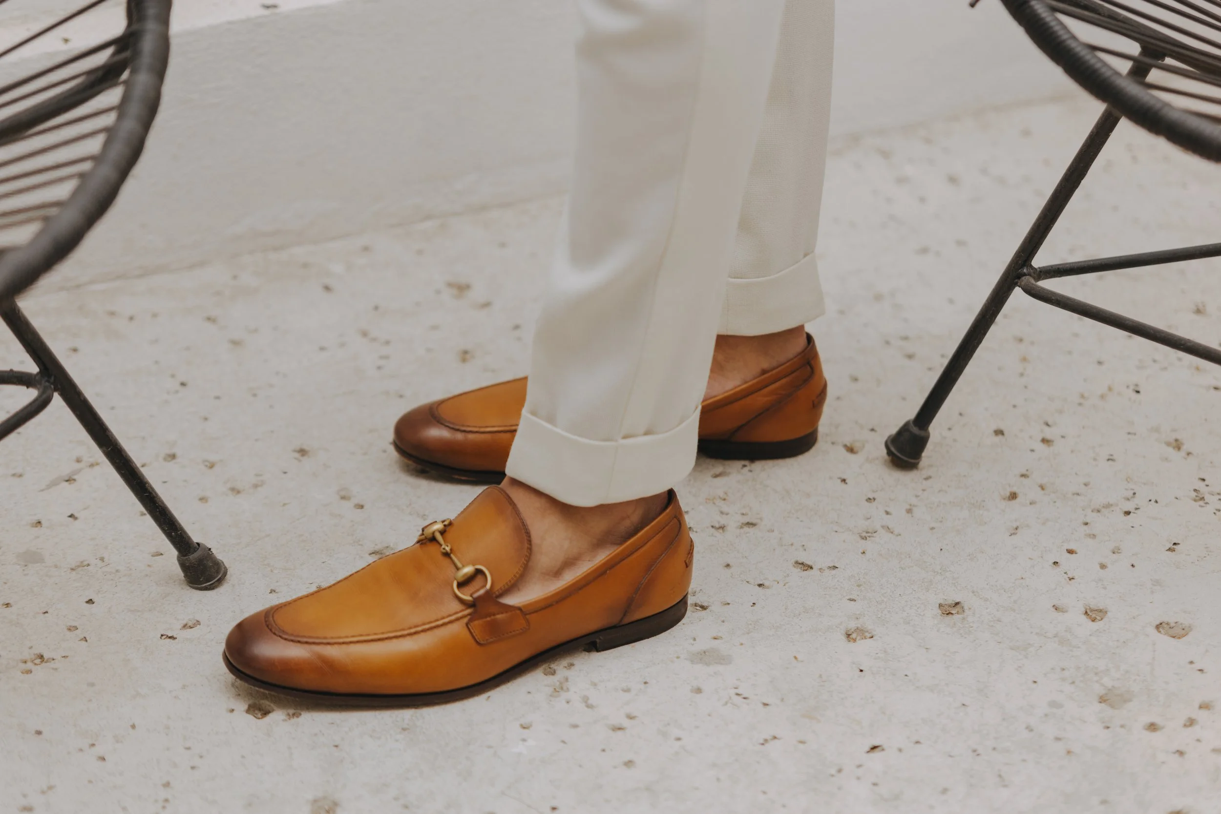 Close-up of a person wearing tan loafers and white pants, sitting at a table on a speckled tile floor.