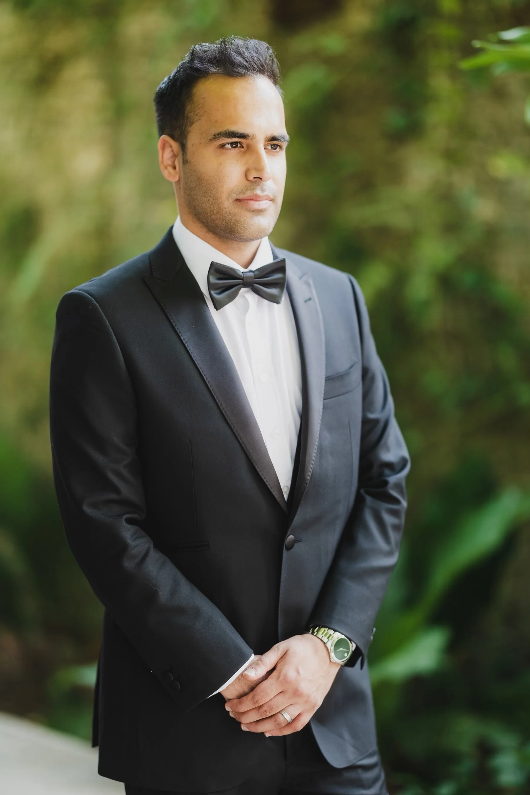 A man dressed in a black tuxedo with a white shirt and black bow tie, standing outdoors with greenery in the background.