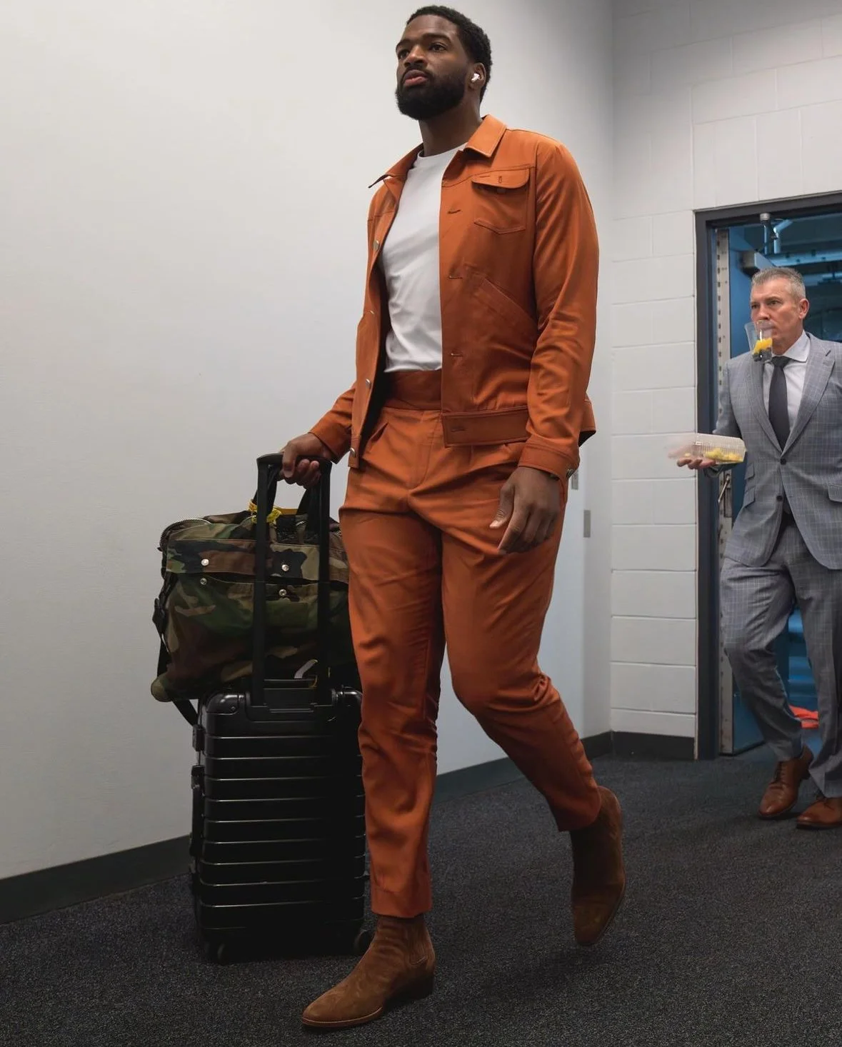 A man in an orange suit walking through an airport with a rolling suitcase and a camouflage duffel bag, and a man in a gray suit in the background holding a tray of food.