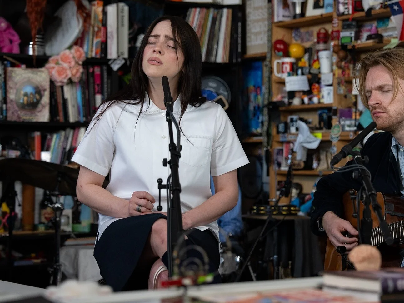 A woman singing and a man playing guitar in a room filled with books and miscellaneous items.