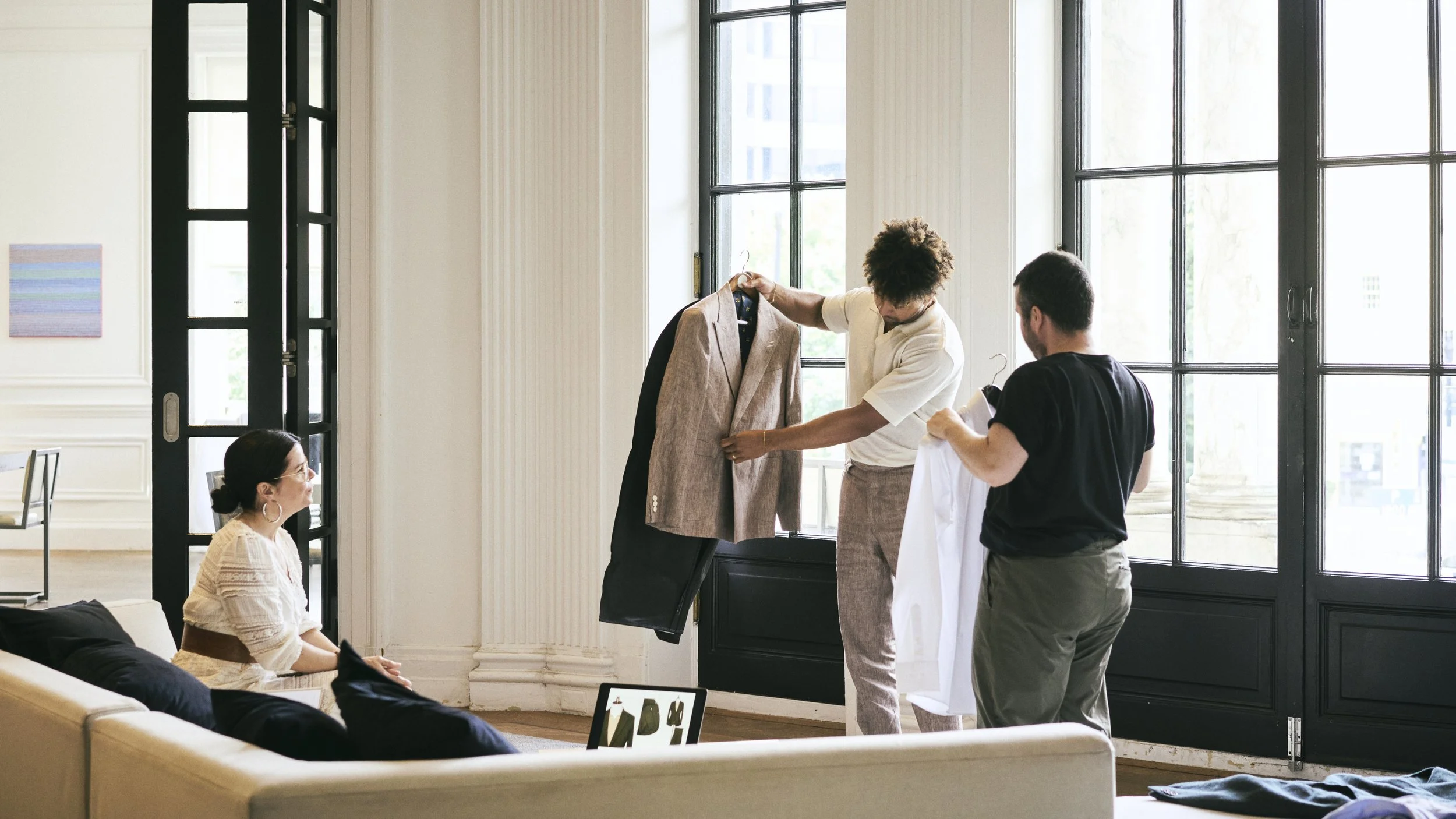 Three people in a bright, modern room with large windows and black doors; two men are holding clothing, one woman is sitting on a beige sofa, looking at them.