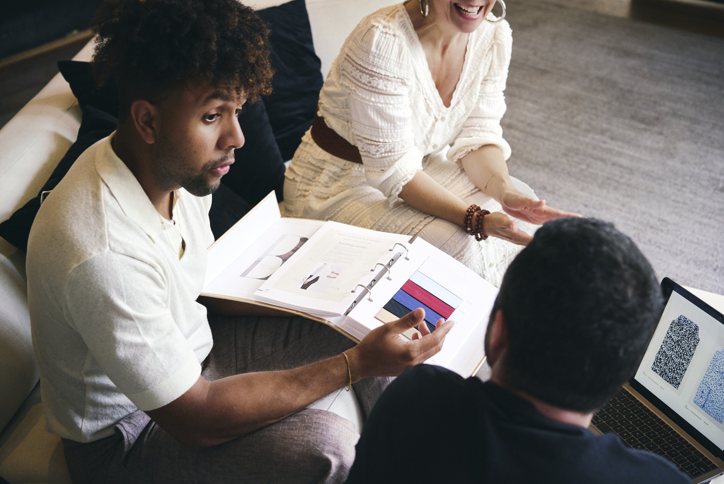 Three people sitting on a beige couch engaged in a discussion, with a laptop displaying QR codes and a binder with fabric color samples and documentation on their laps.