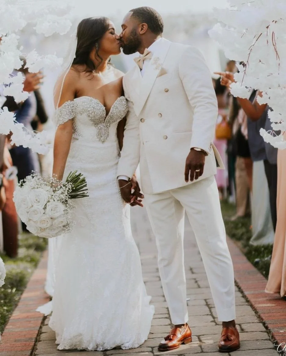 A bride and groom share a kiss during their wedding ceremony in an outdoor setting. The bride wears a white lace wedding dress with off-the-shoulder sleeves and holds a bouquet of white roses. The groom wears a white double-breasted suit with brown s
