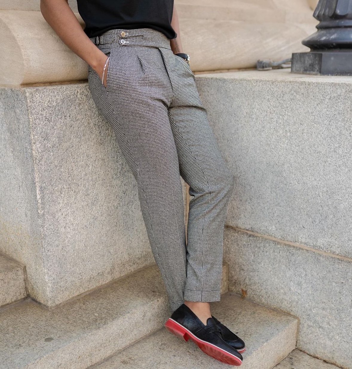 Person wearing black loafer shoes with red soles, checkered pants, black shirt, standing against stone steps and wall.