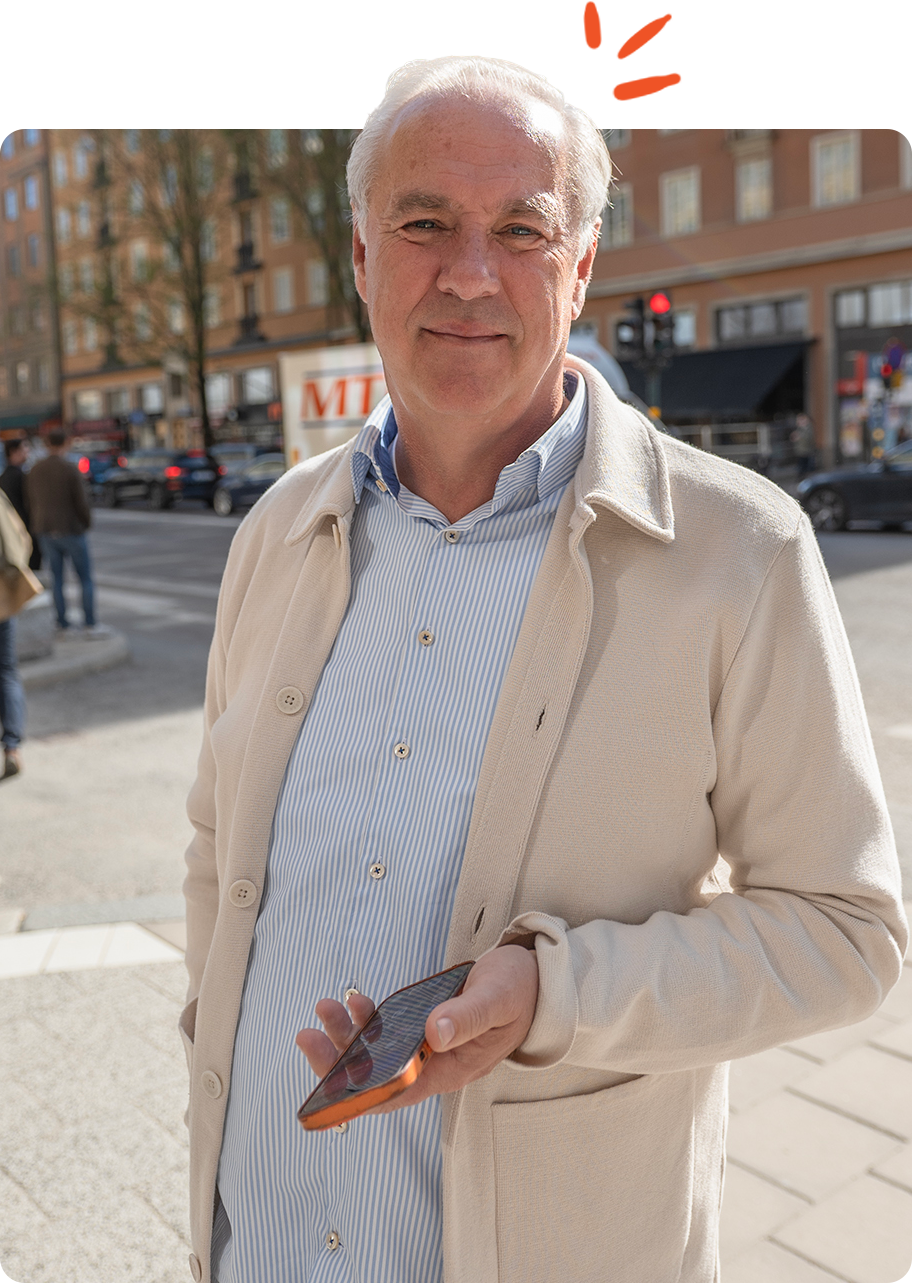 En äldre man står utomhus i ett stadsområde, bär en ljusfärgad kappa och en rutig skjorta, håller i en mobiltelefon och ler lätt mot kameran.