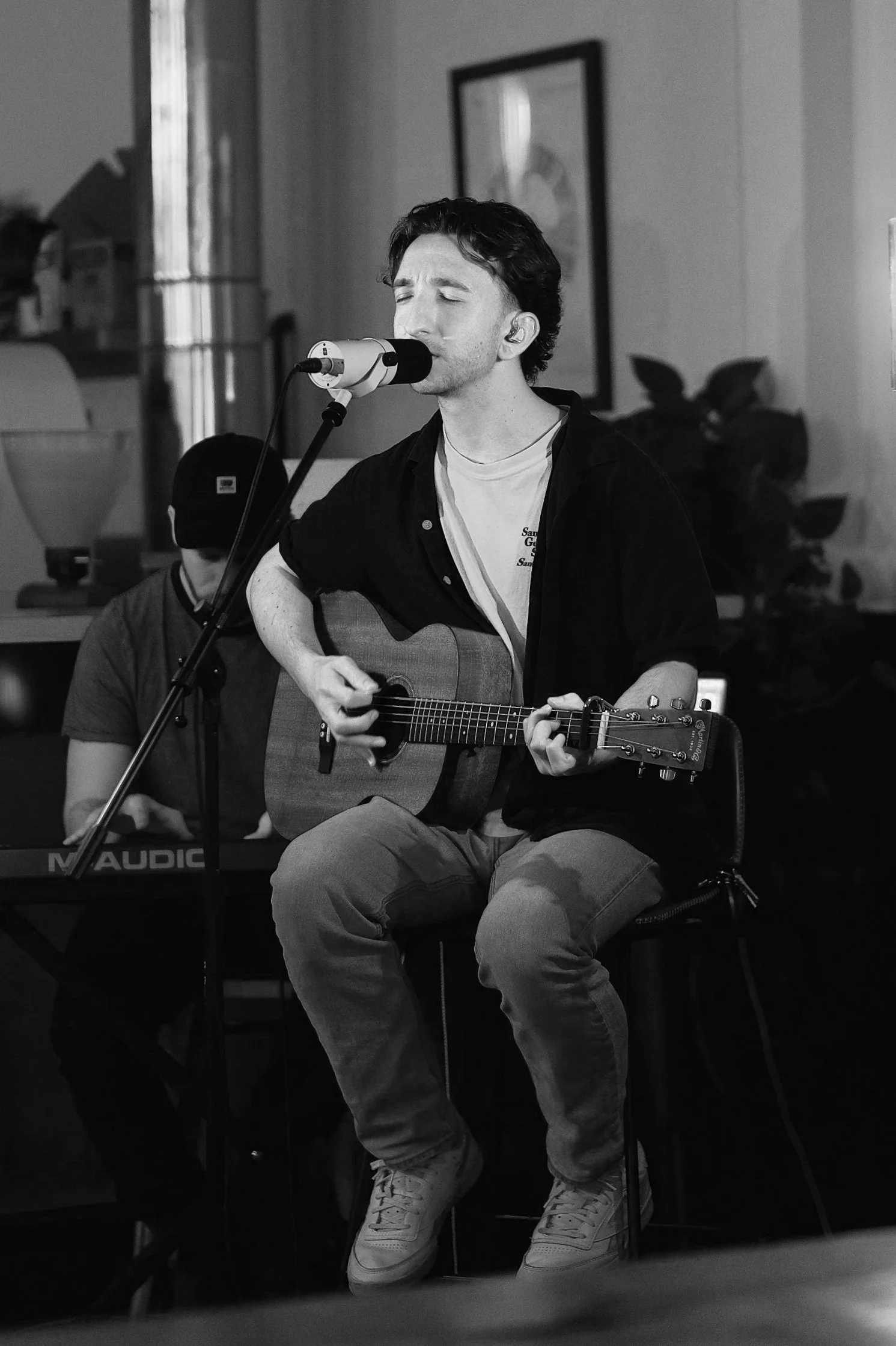 A man with dark curly hair closed eyes, singing into a microphone, playing an acoustic guitar, seated, with a keyboardist in the background in an indoor setting.