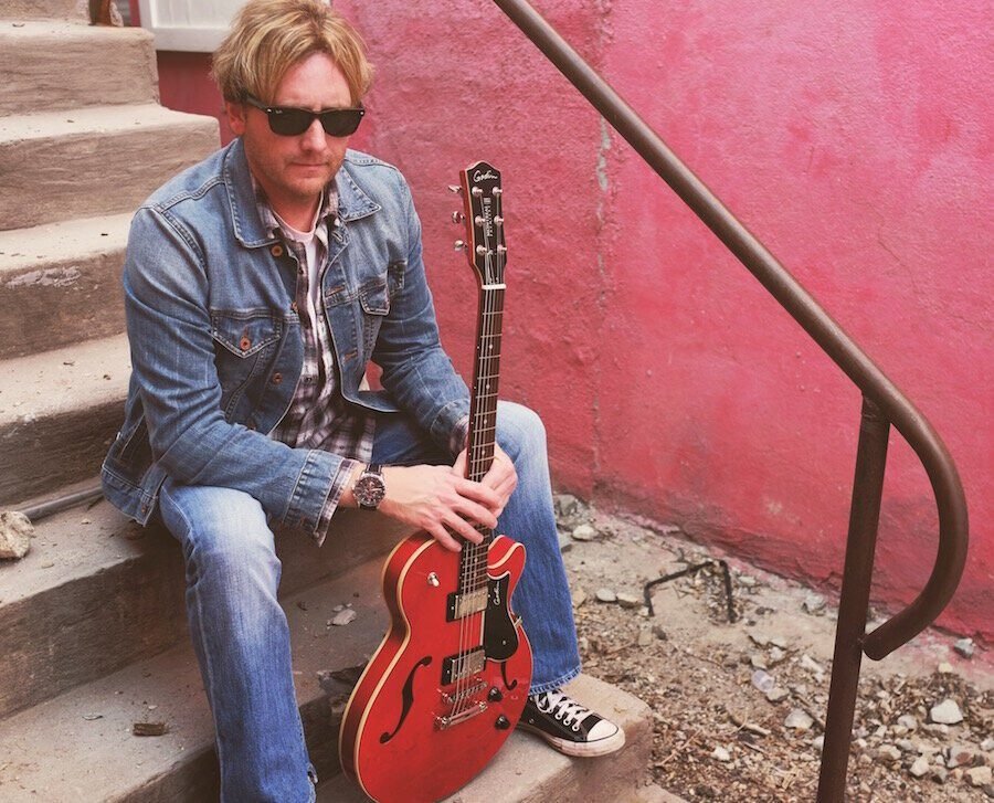 American singer-songwriter Scott Helmer seated on outdoor steps with a red electric guitar during a promotional photo session.