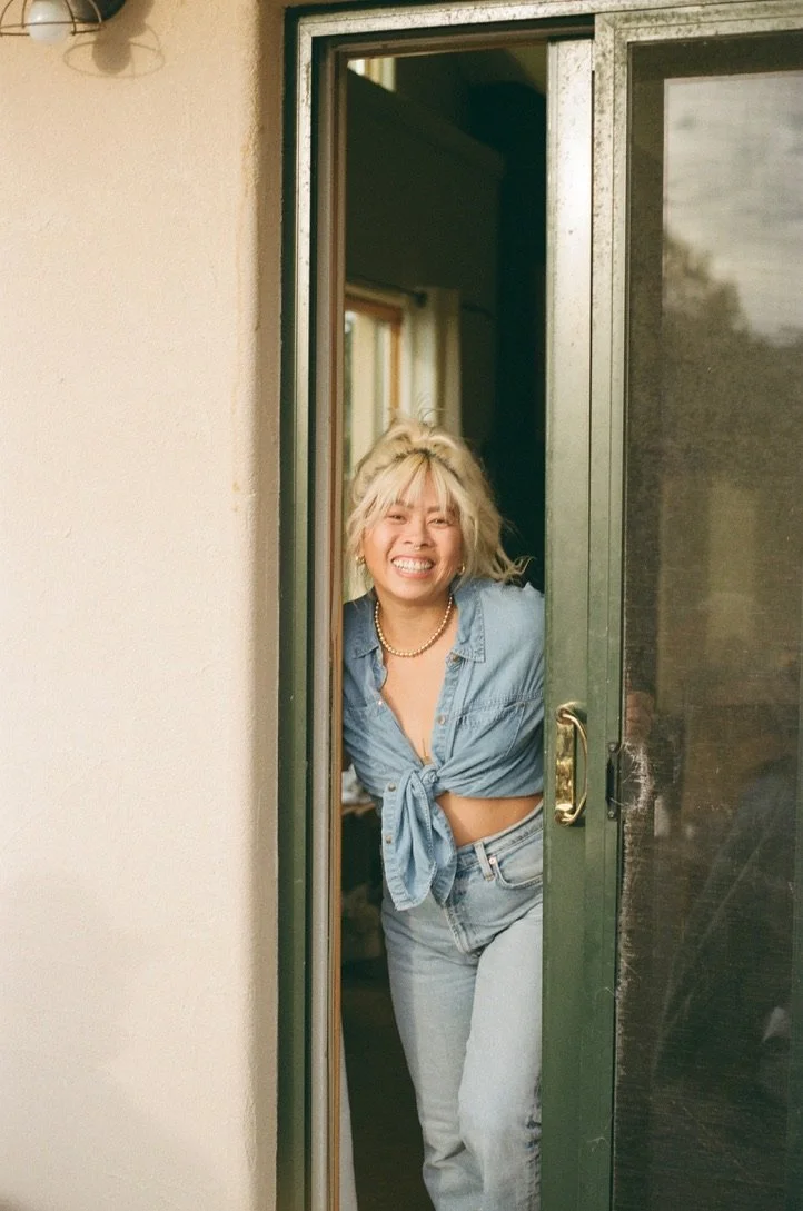 A woman with blonde curly hair smiling and leaning through a doorway, wearing a tied denim shirt and jeans.