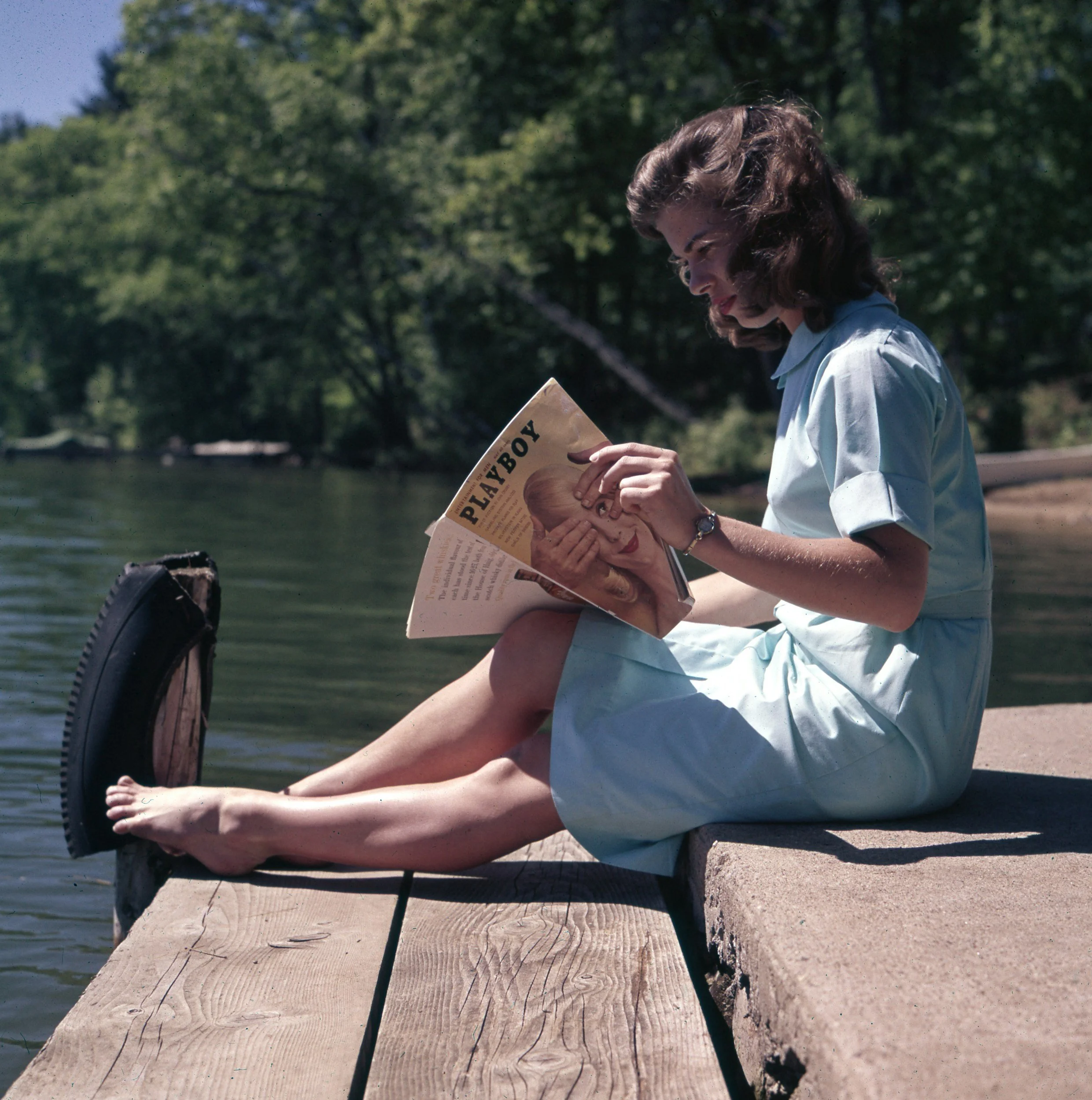 A woman sitting on a wooden dock by the water, reading a vintage Playboy magazine with her legs with toes in the water, surrounded by trees.