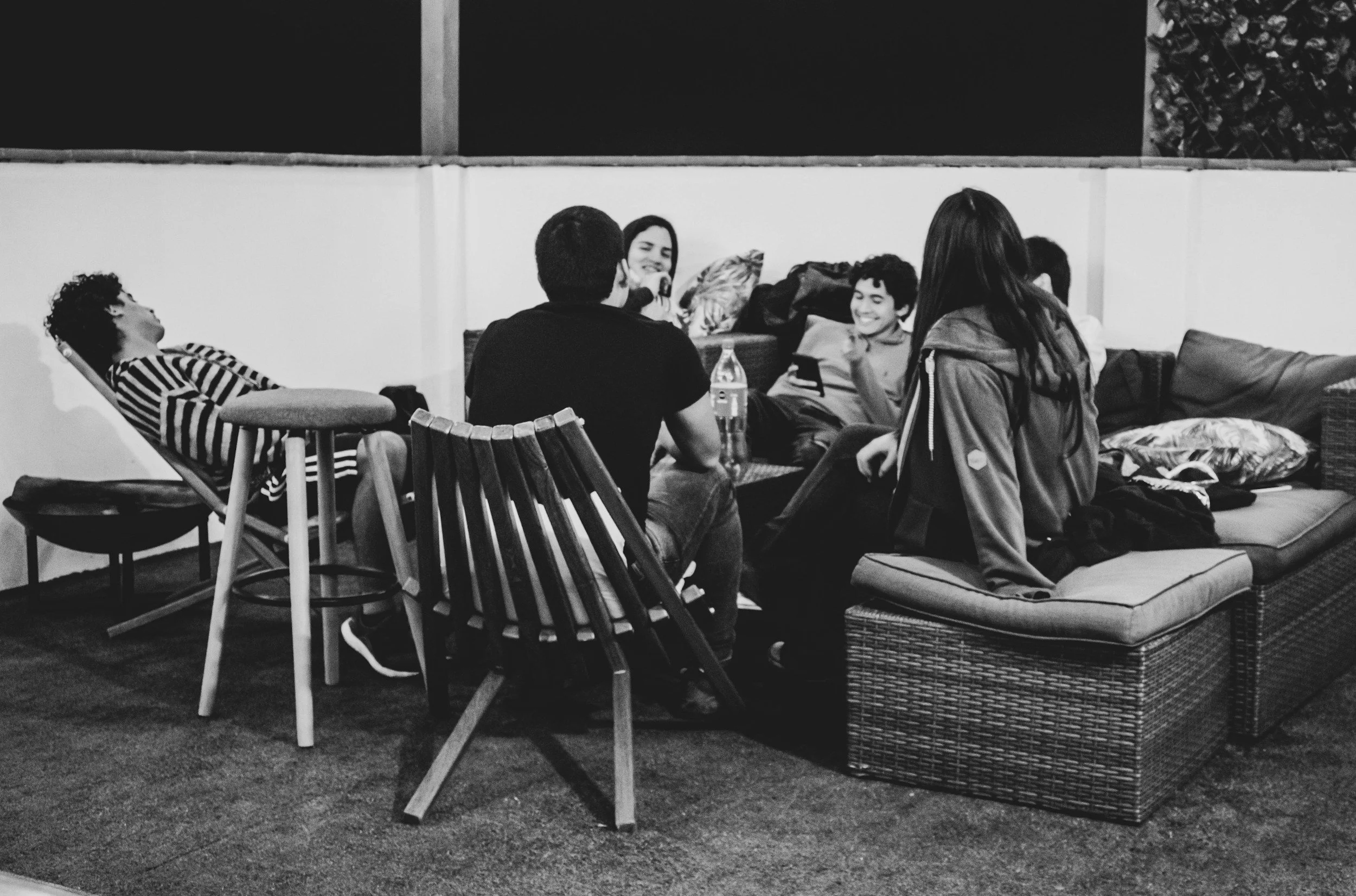 A group of six friends sitting and chatting on outdoor patio furniture, some laughing and smiling, with a bottle of water on the table.