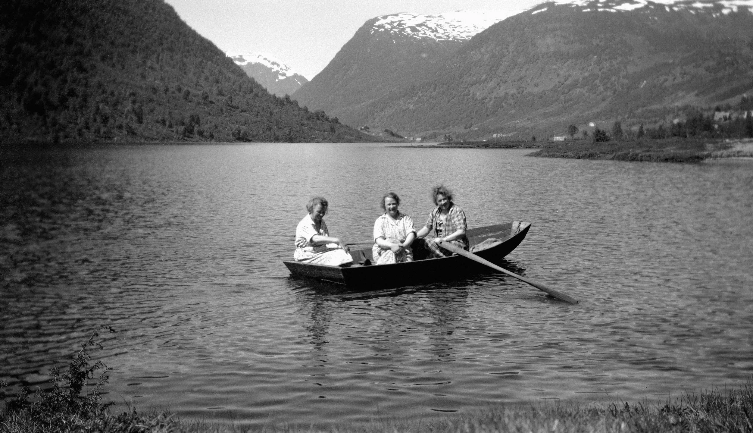 Three women sitting in a small boat on a calm lake surrounded by mountains.